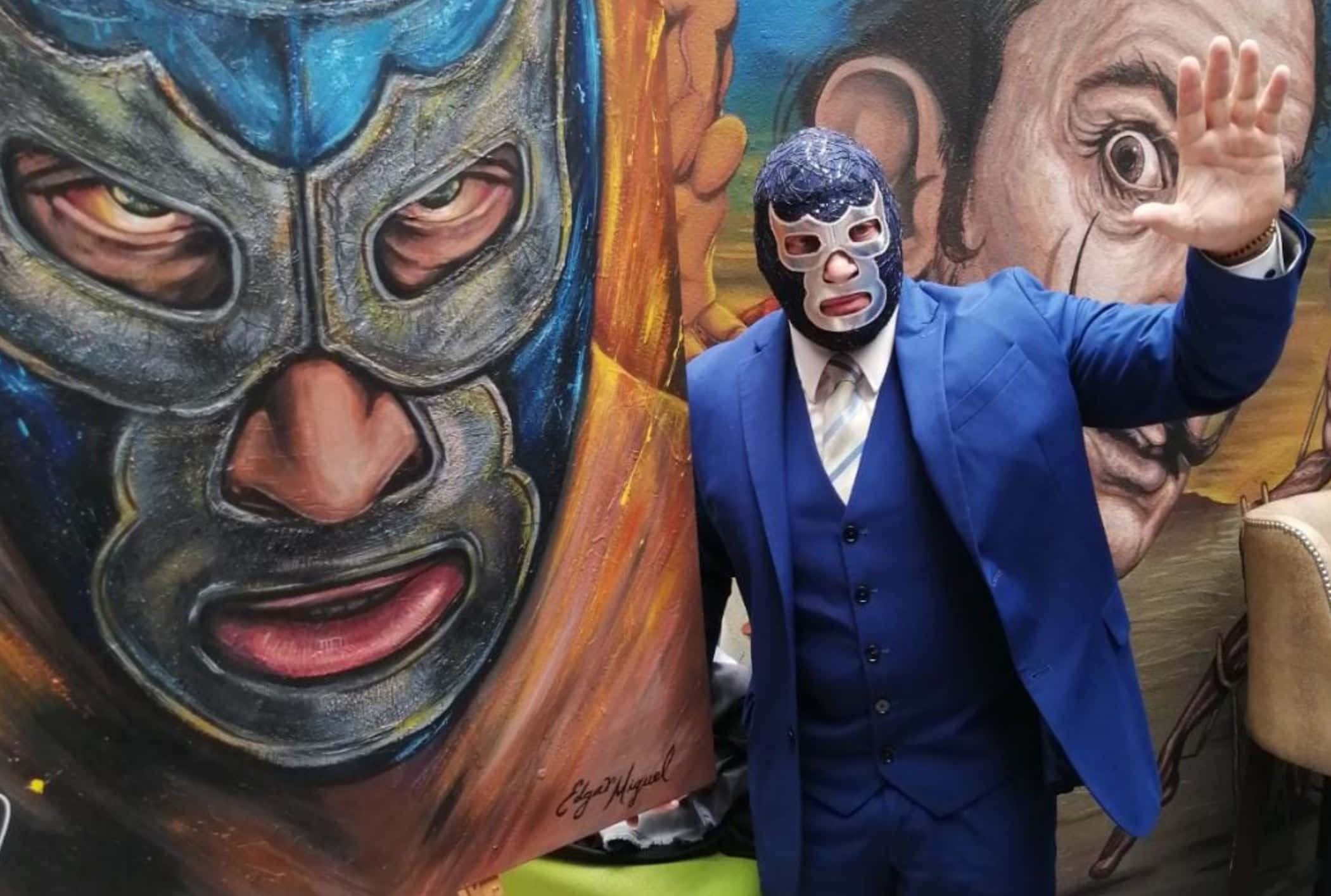 Mexican 桌布 壁紙 — 一名身穿藍色西裝、戴著自由摔角面具的男子，在一幅大型戴面具摔角手壁畫旁揮手。