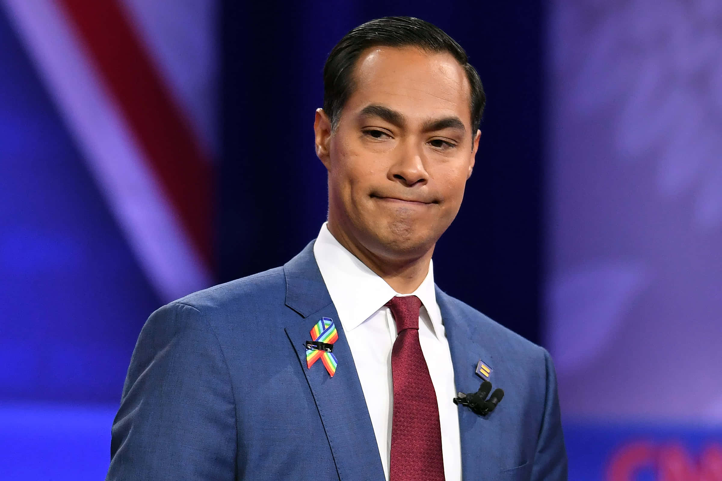 Celebrity 桌布 壁紙 — 一名身穿藍色西裝、紅色領帶的男子，在模糊的愛國背景舞台上，佩戴彩虹和旗幟別針，露出微妙微笑。