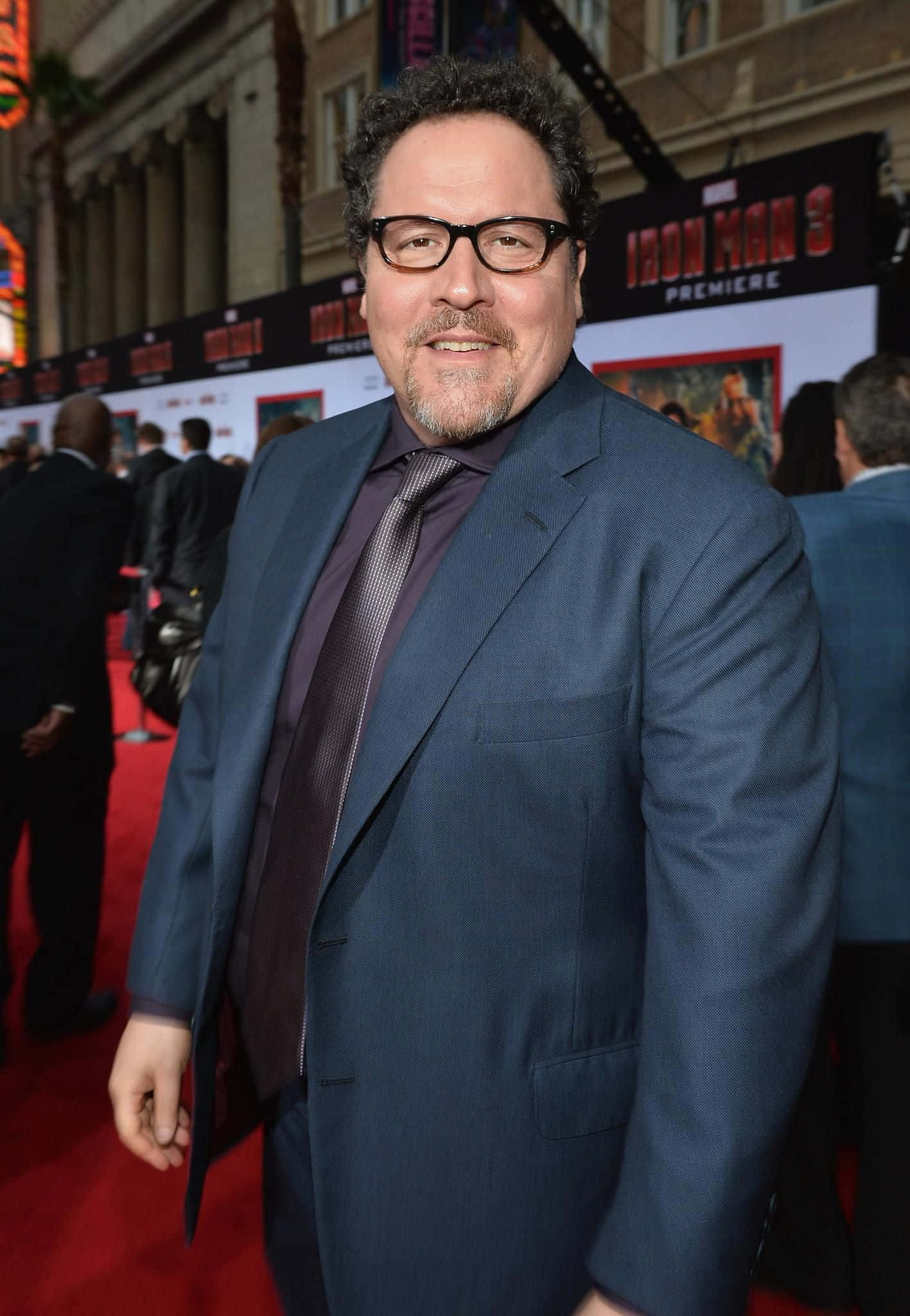 Jon Favreau 桌布 壁紙 — 一名身穿藍色西裝及戴眼鏡的男子在《鋼鐵人3》首映會的紅地毯上微笑，身後有電影海報。
