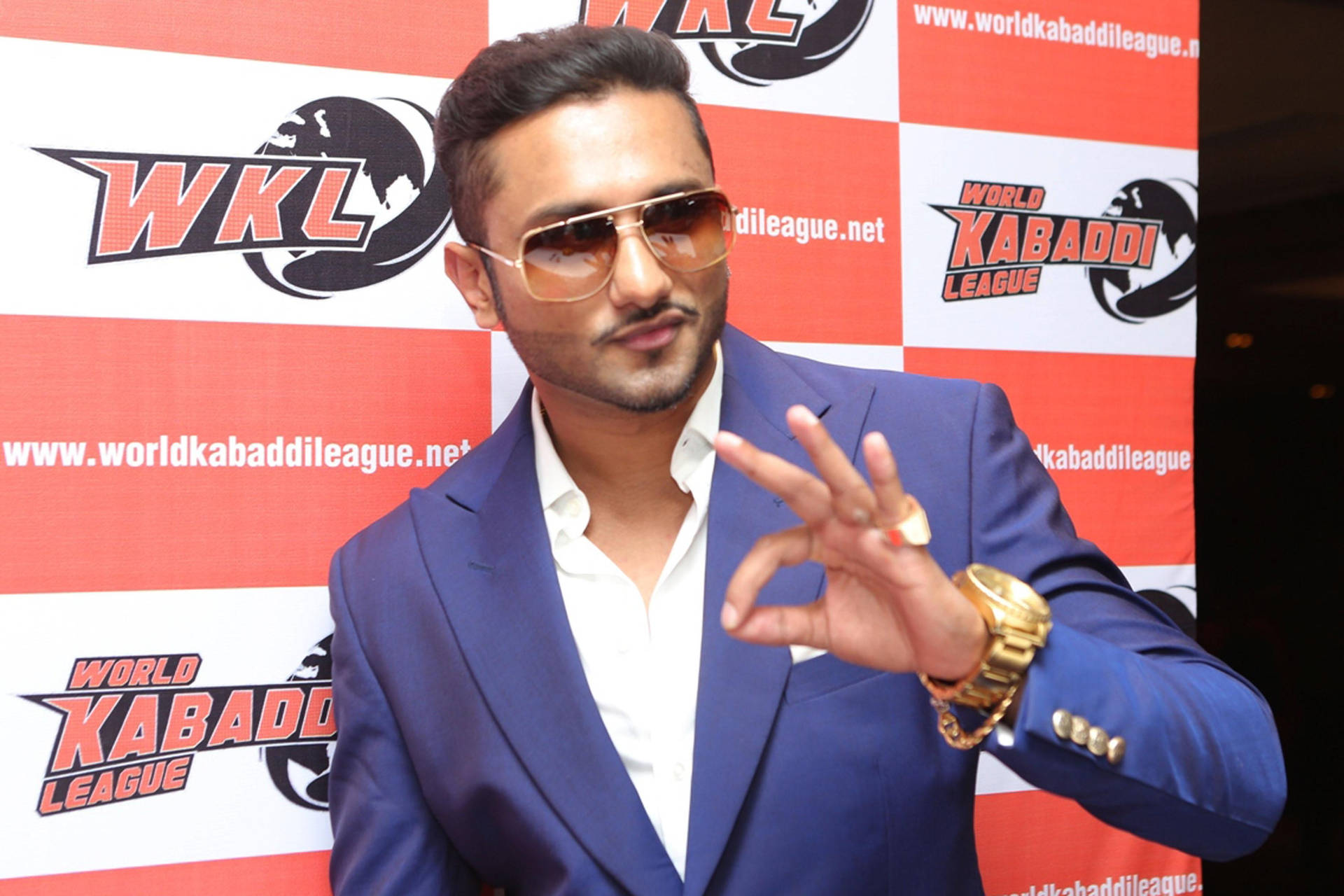  壁紙 — 一名身穿藍色西裝和金色太陽眼鏡的男子，在紅白相間的World Kabaddi League背景前做出OK手勢。