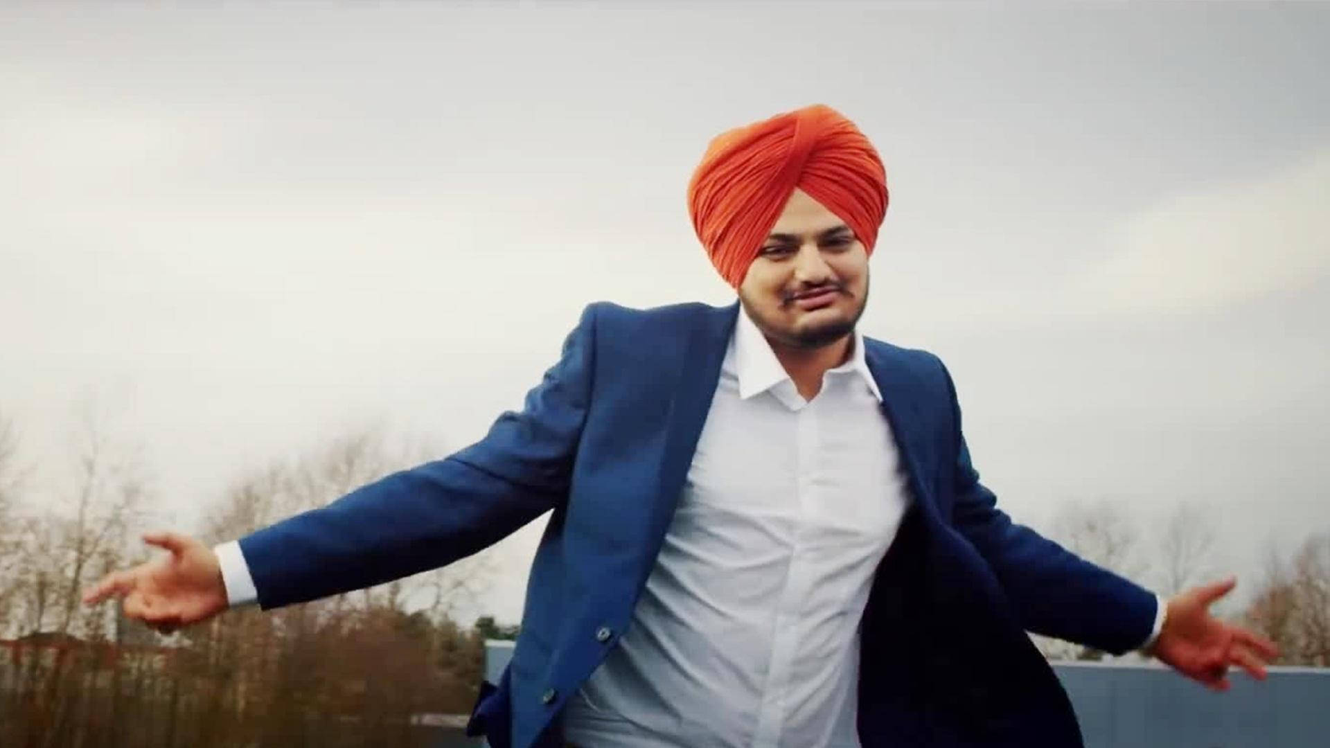 身穿藍色西裝外套和橙色頭巾的男子，在戶外張開雙臂站立。 — Sidhu Moose Wala 壁紙