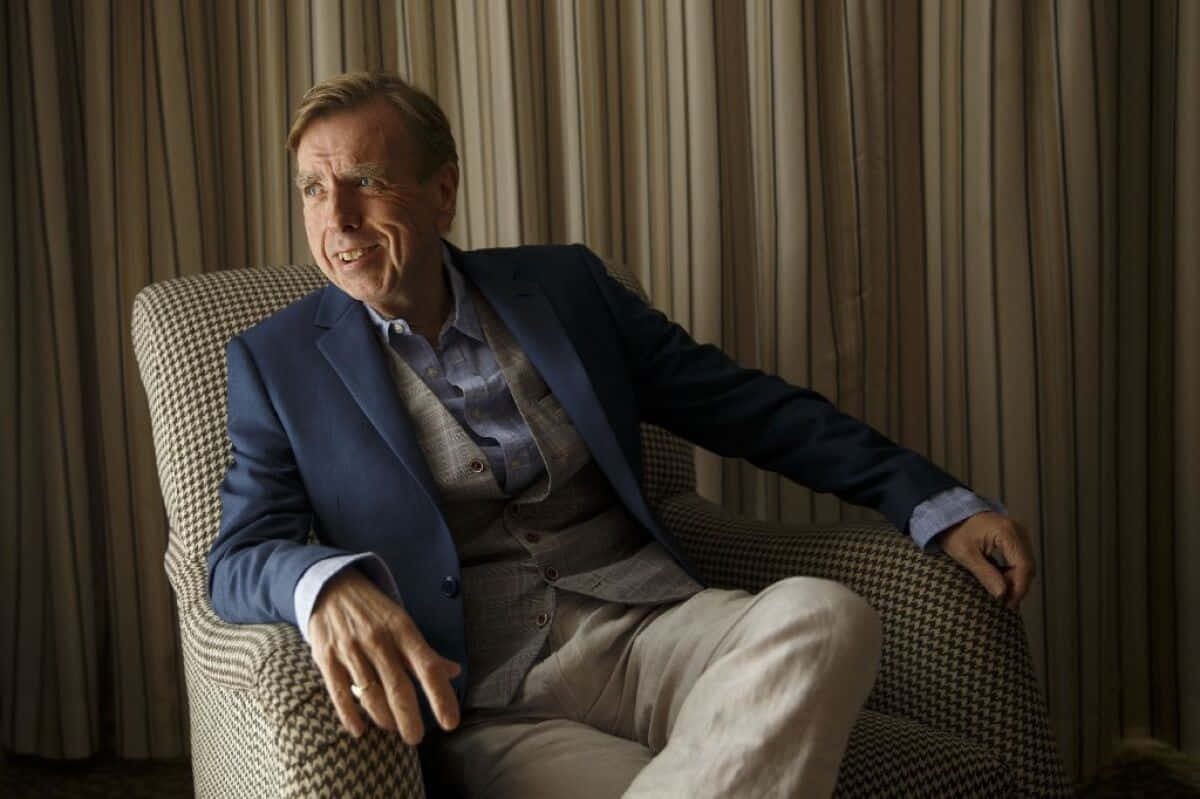 Timothy Spall 桌布 壁紙 — 一名身穿藍色西裝外套和米色長褲的男子，放鬆地坐在一張千鳥格扶手椅上，背景是條紋窗簾。