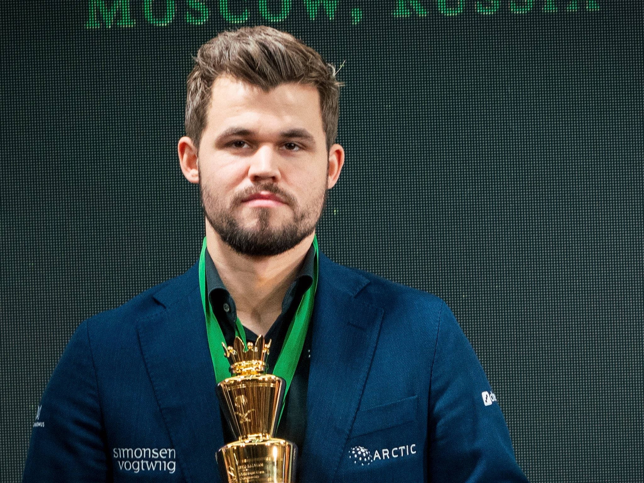 Magnus 桌布 壁紙 — 一名身穿藍色西裝外套的男子，在顯示「MOSCOW RUSSIA」的深綠色數位螢幕前手持金色獎盃。