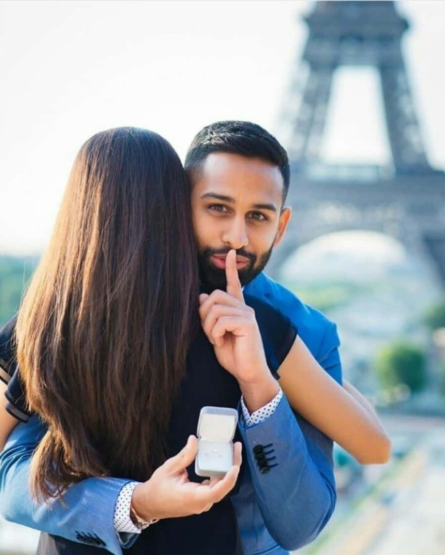 Proposal 桌布 壁紙 — 一名身穿藍色西裝的男子向驚訝的伴侶低聲說出秘密，伴侶手持打開的戒指盒，背景是艾菲爾鐵塔。