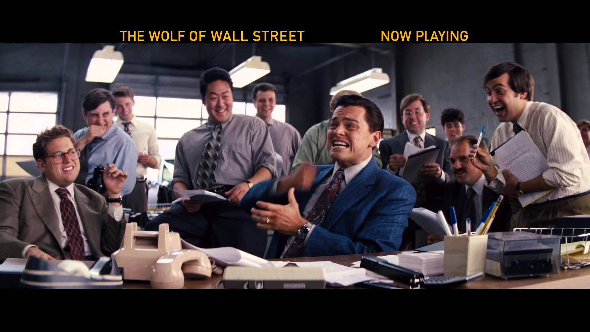 Wolf Of Wall Street 桌布 壁紙 — 一名身穿藍色西裝的男子在他凌亂的辦公桌上狂笑，同事們在混亂的辦公室裡圍著他歡呼。
