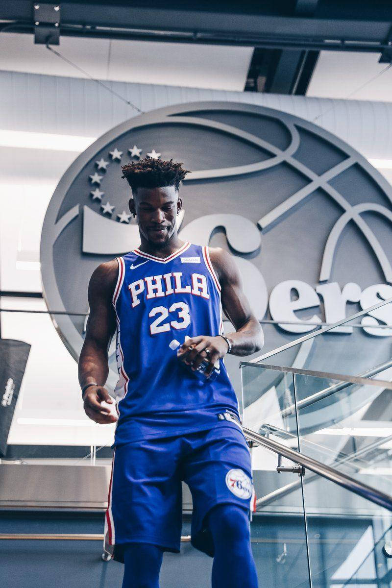 NBA 桌布 壁紙 — 一名身穿藍色費城76人球衣的籃球運動員自信地站在大型球隊標誌牆前。