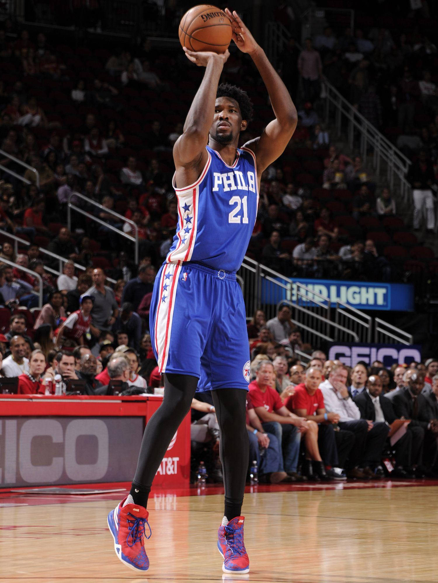 Embiid 桌布 壁紙 — 一名身穿藍色費城76人球衣的籃球運動員在燈光明亮的場館中跳起投籃，場館內座無虛席。