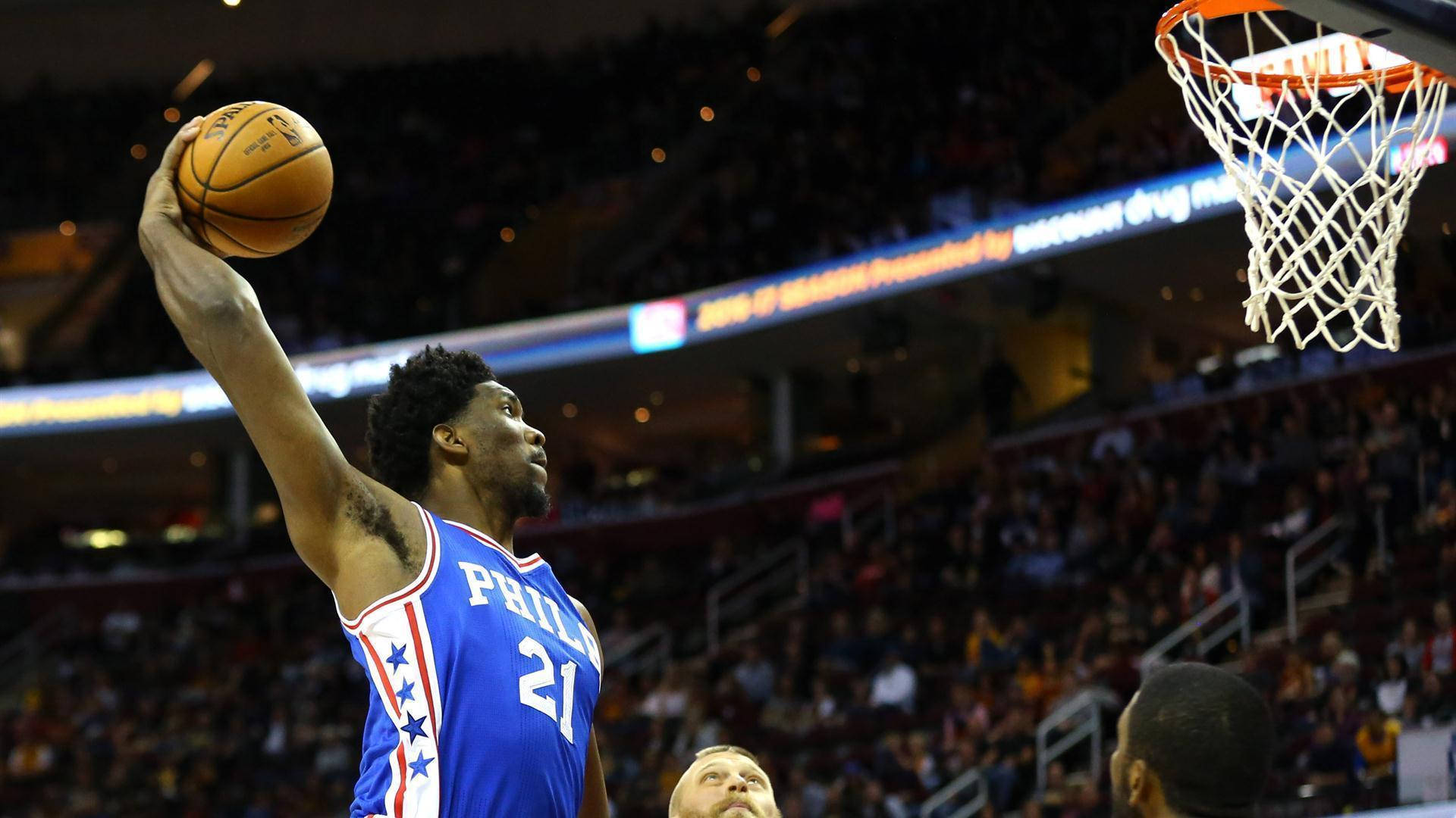 Embiid 桌布 壁紙 — 一名身穿藍色費城球衣的籃球運動員在明亮的燈光下，在擠滿觀眾的場館中高高躍起灌籃。