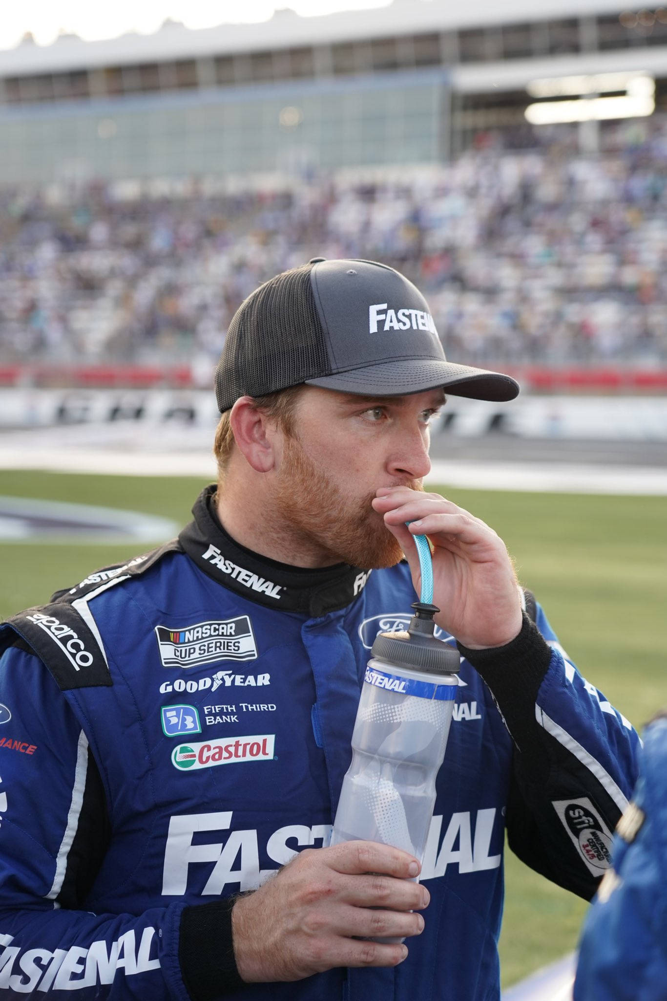 Drinking 桌布 壁紙 — 一名身穿藍色賽車服的NASCAR車手在賽道上從瓶子裡喝水，背景是模糊的人群。