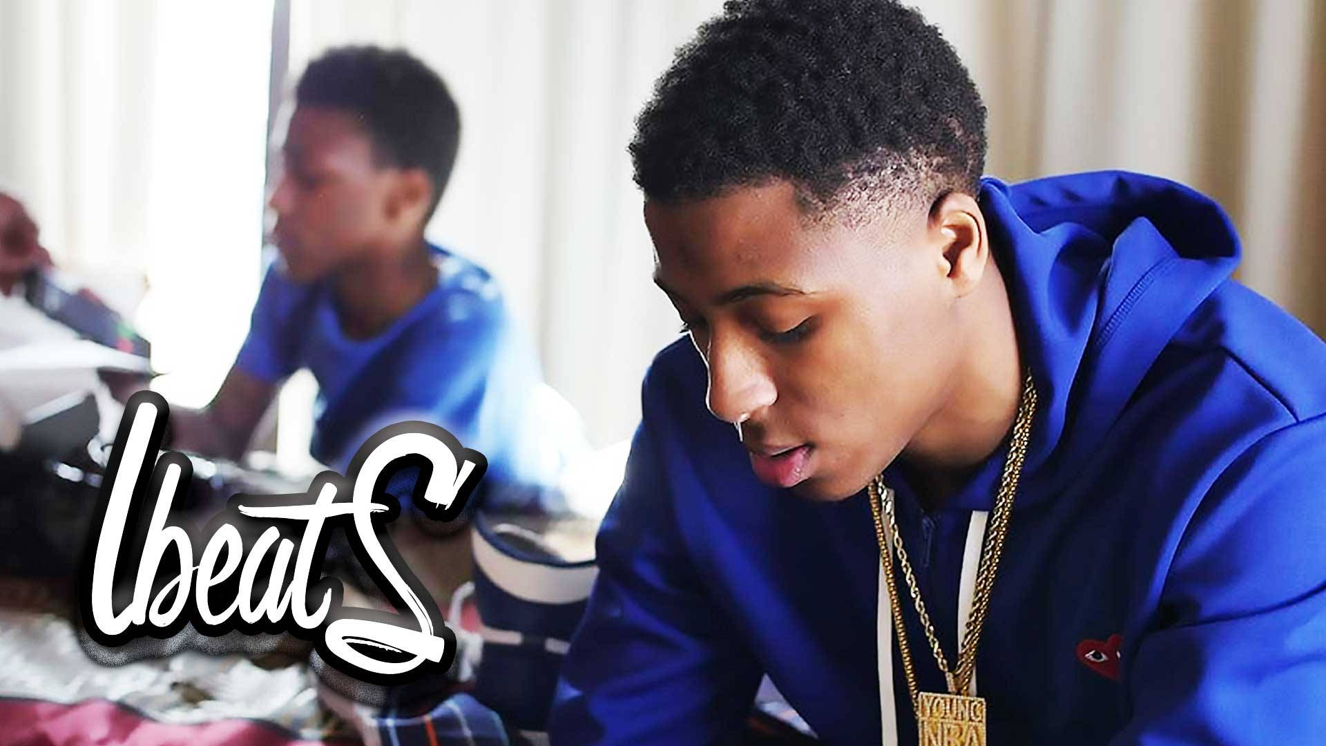 Youngboy 桌布 壁紙 — 一名身穿藍色連帽衫、戴著金鏈的年輕男子專注地向下看，背景中另一個人模糊地坐著。