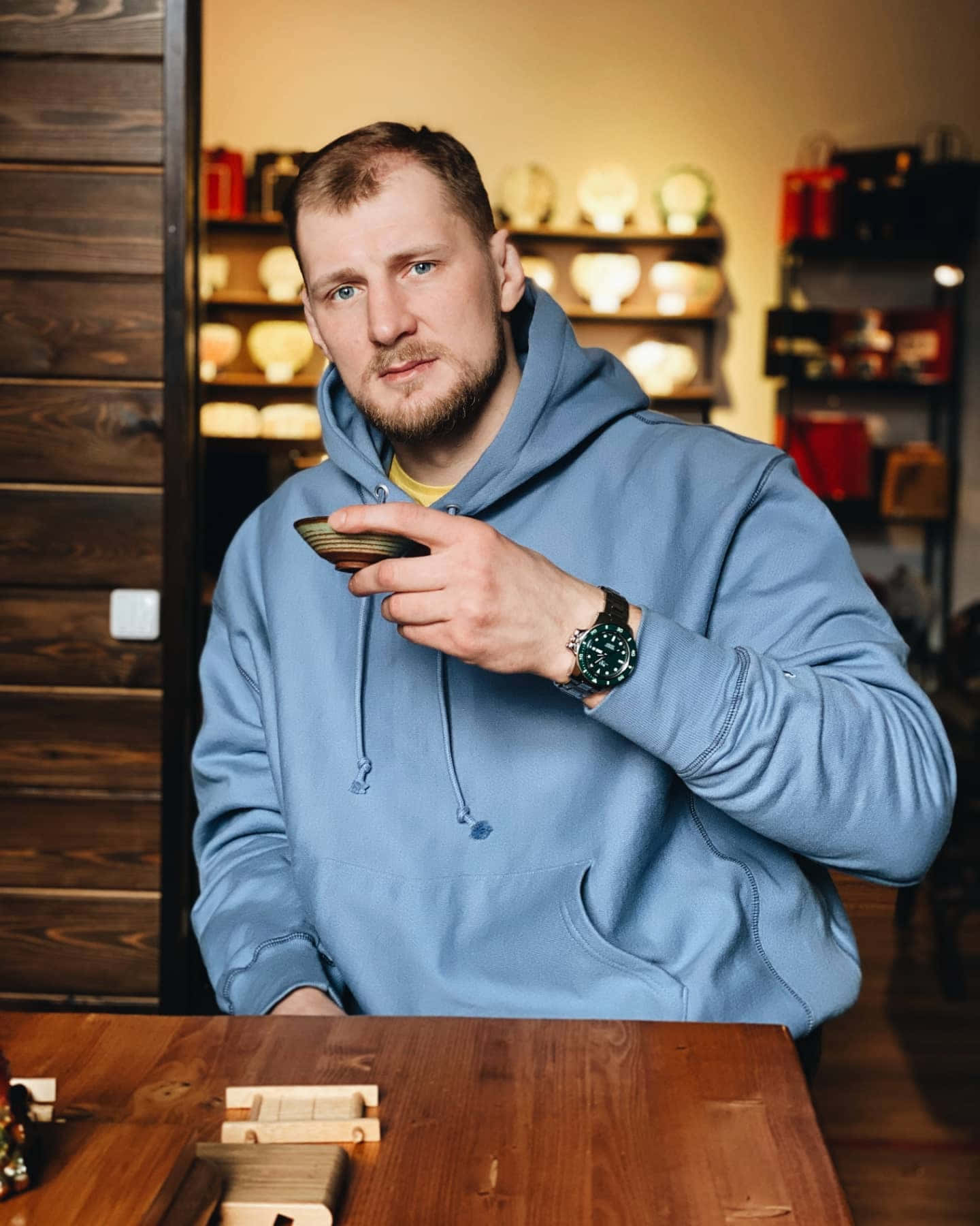 Celebrity 桌布 壁紙 — 一名身穿藍色連帽衫的男子坐在木桌旁，身後是溫暖照明的房間，架子上擺滿陶器，他手持一個小碗。