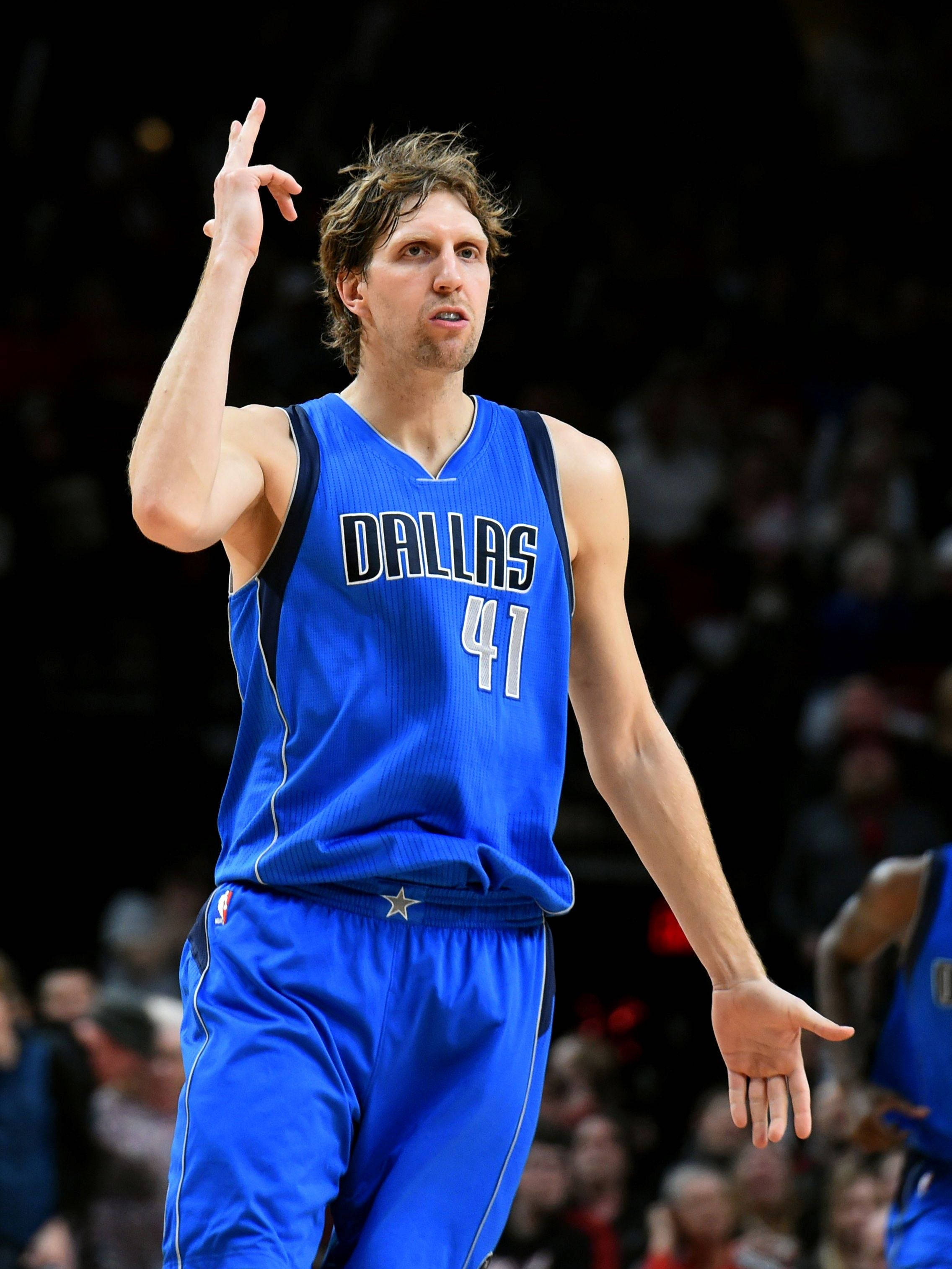Dirk Nowitzki 桌布 壁紙 — 一名身穿藍色達拉斯球衣41號的籃球運動員在明亮的場館燈光下比賽期間舉起右手做手勢。