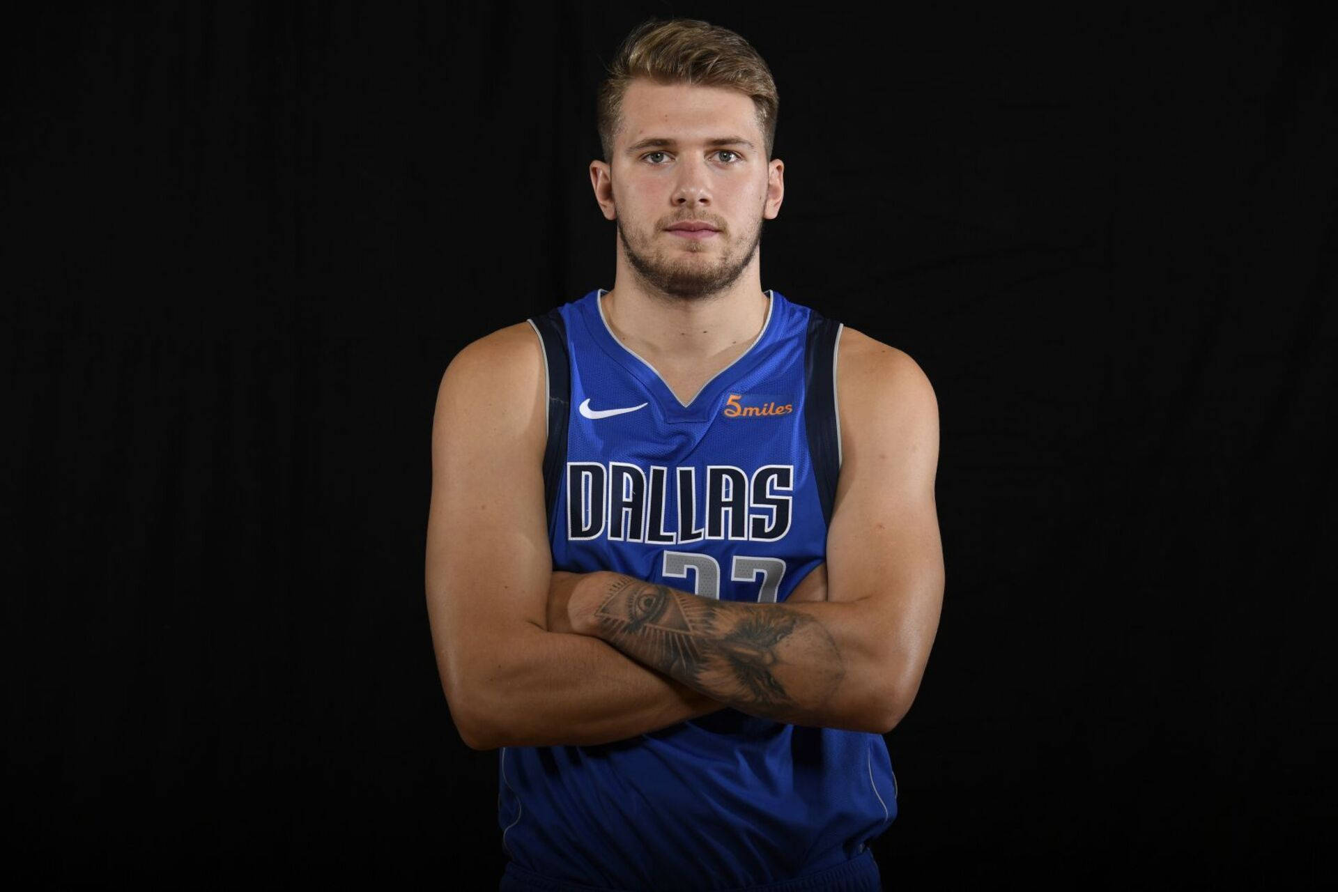 Luka 桌布 壁紙 — 一名身穿藍色達拉斯球衣的籃球運動員自信地交叉雙臂，站在黑色背景前。