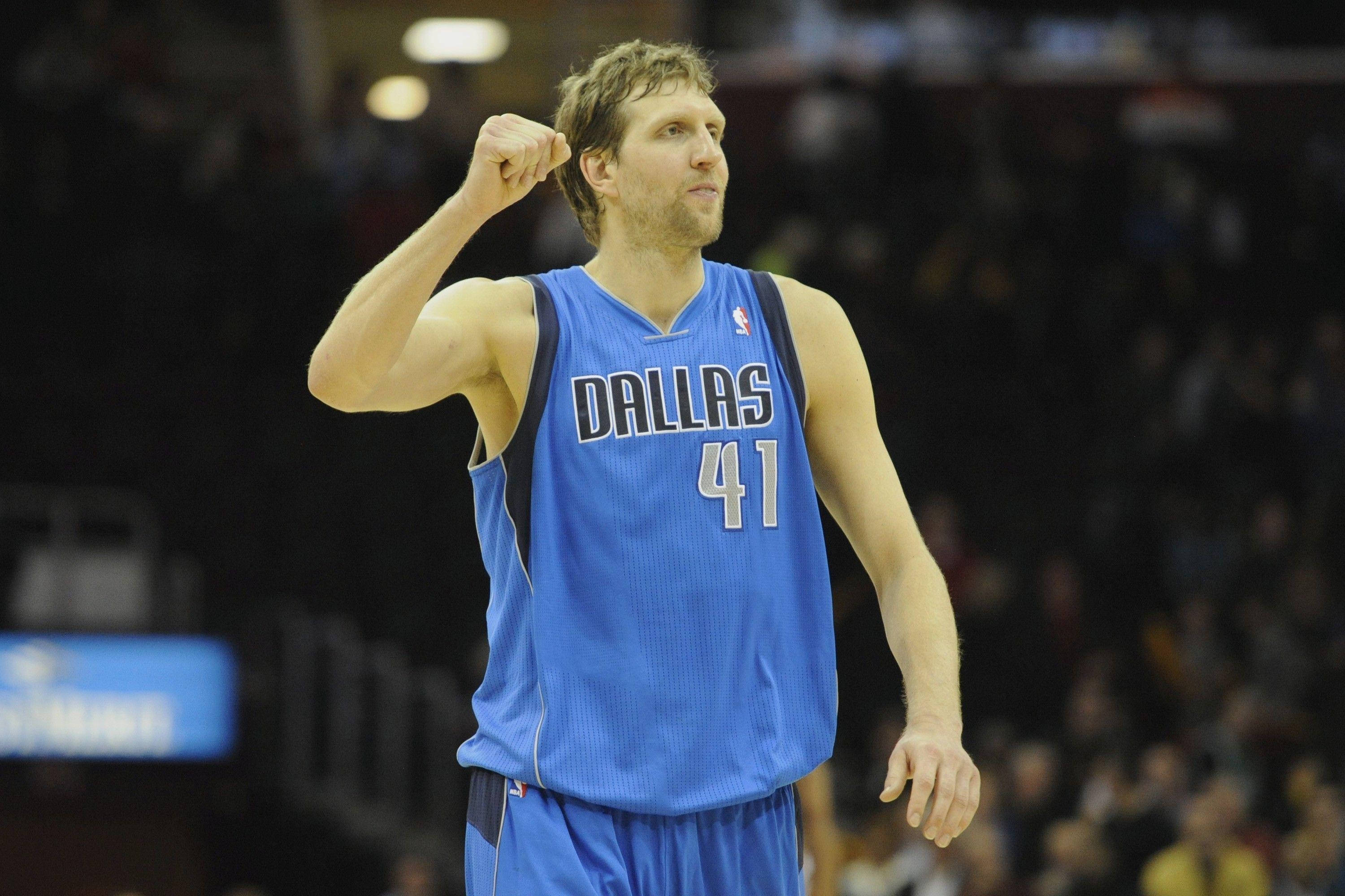 Dirk Nowitzki 桌布 壁紙 — 一名身穿藍色達拉斯球衣的籃球運動員在場館燈光下的比賽中高舉拳頭慶祝勝利。