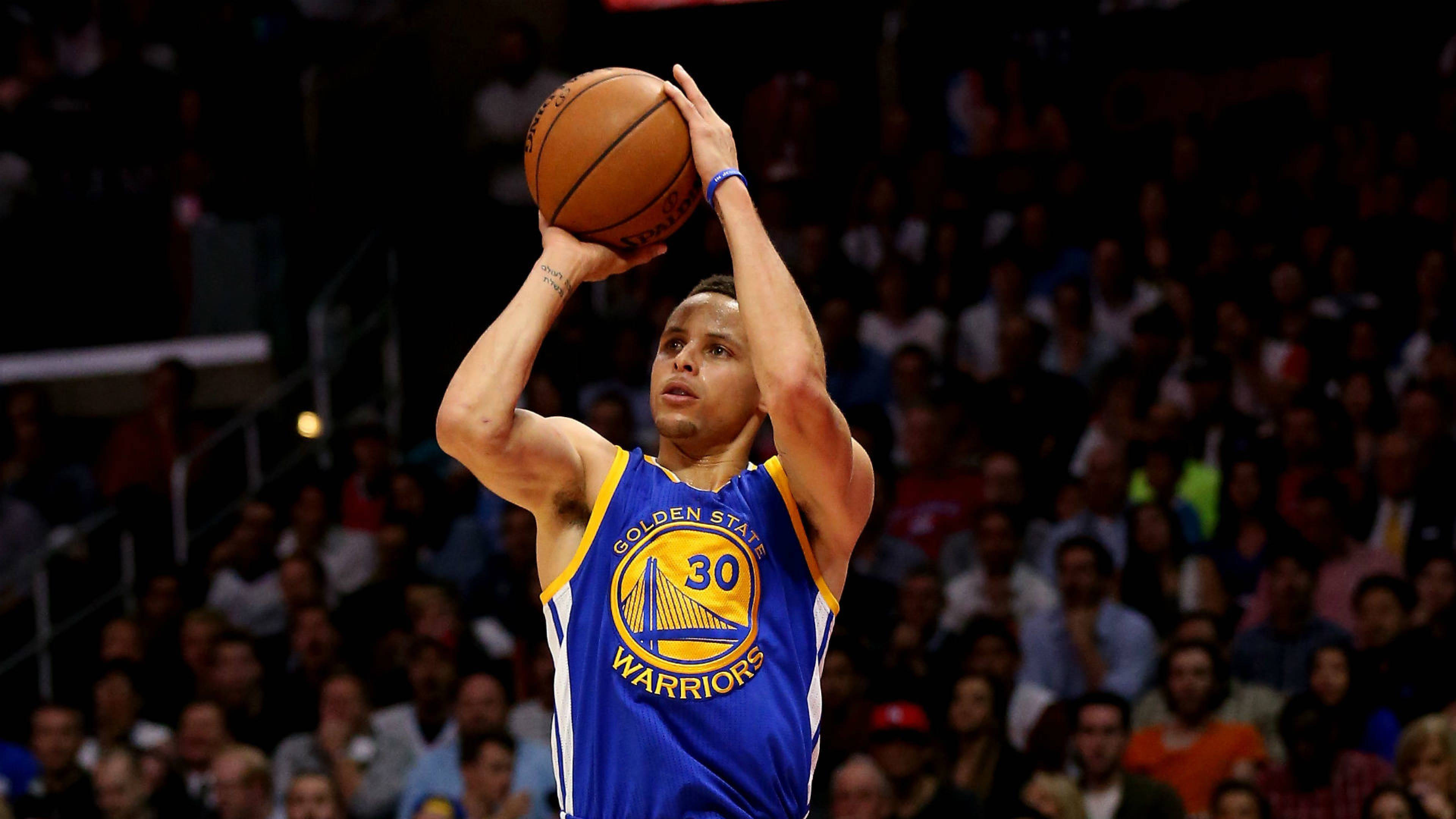 4K NBA 桌布 壁紙 — 一名身穿藍色金州勇士隊球衣的籃球運動員，在明亮的場館燈光下準備投籃，身後是模糊的人群。