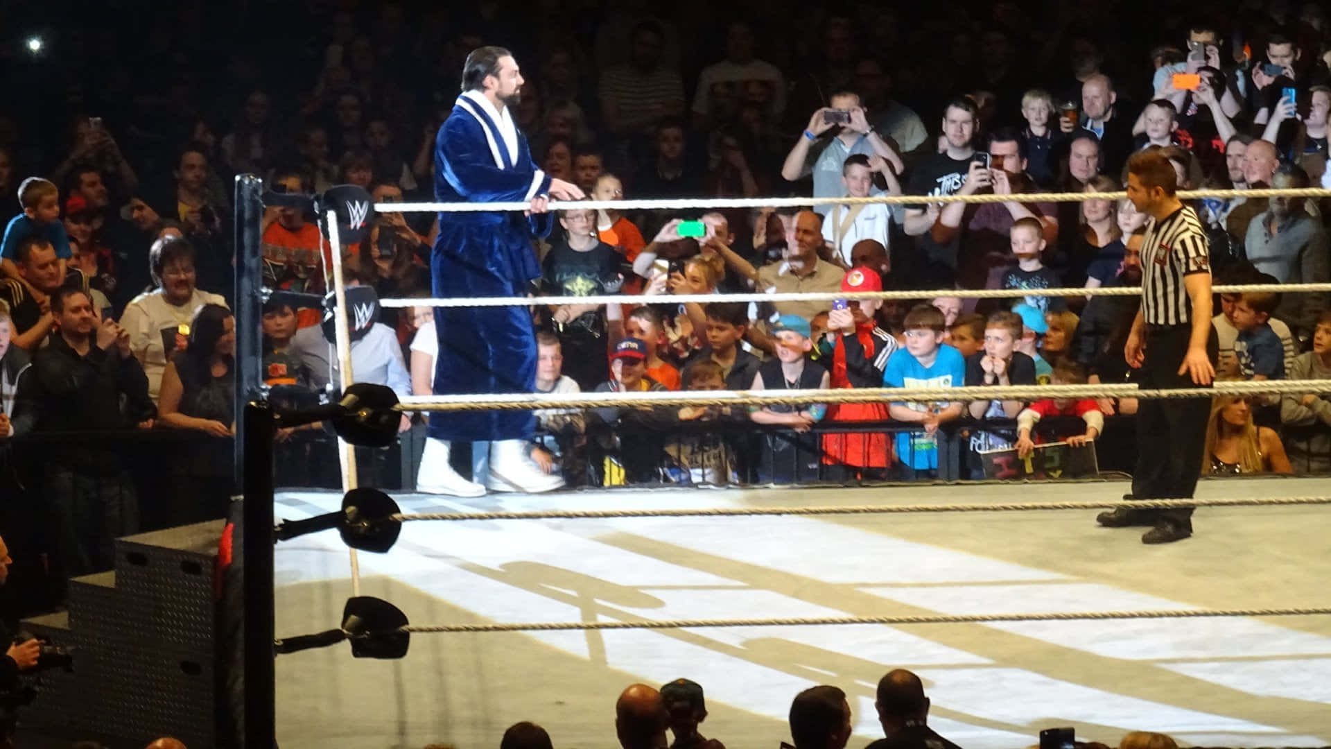WWE 桌布 壁紙 — 一名身穿藍色長袍的摔角手站在摔角擂台頂部，裁判在旁觀看，粉絲在黑暗的場館中歡呼。