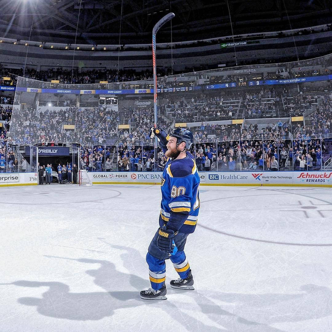 hockey 桌布 壁紙 — 一名身穿藍金球衣的冰球運動員在冰上高舉球棍慶祝勝利，觀眾在玻璃屏障後歡呼。