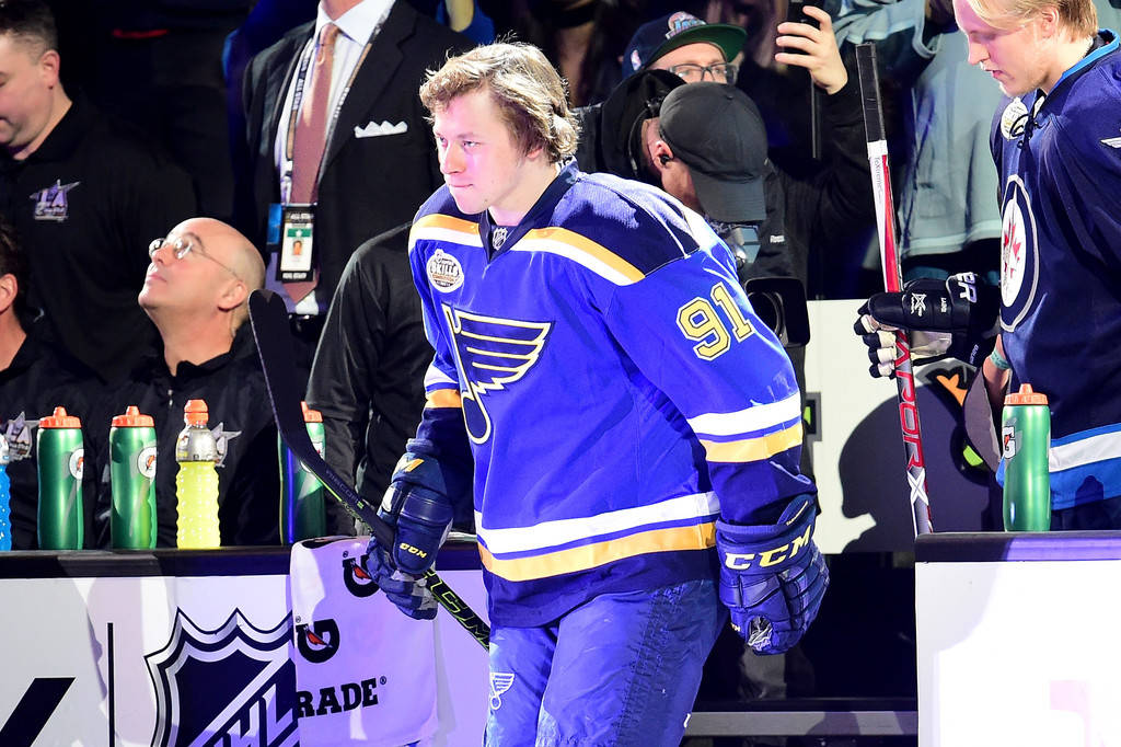 St Louis Blues 桌布 壁紙 — 一名身穿藍金球衣的聖路易斯藍調冰球球員手持球棍步離冰面，背景有隊友和工作人員。