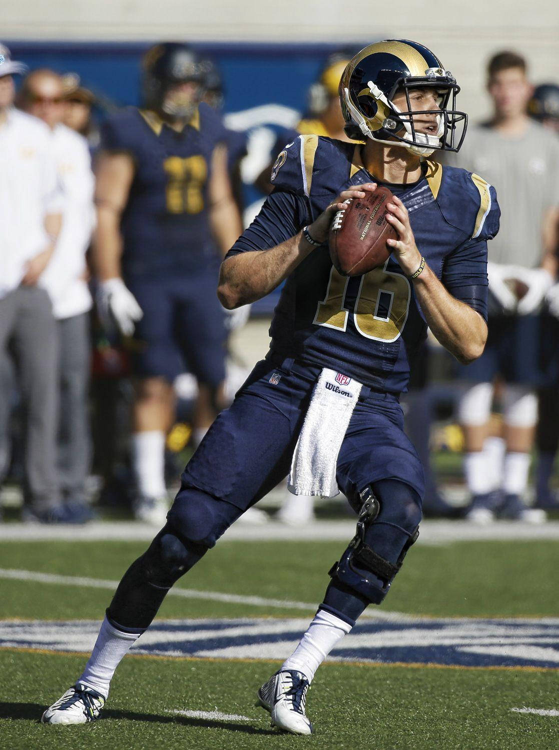 Jared Goff 桌布 壁紙 — 一名身穿藍金色球衣的美式足球四分衛在陽光明媚的球場上，背景有隊友，準備擲球。