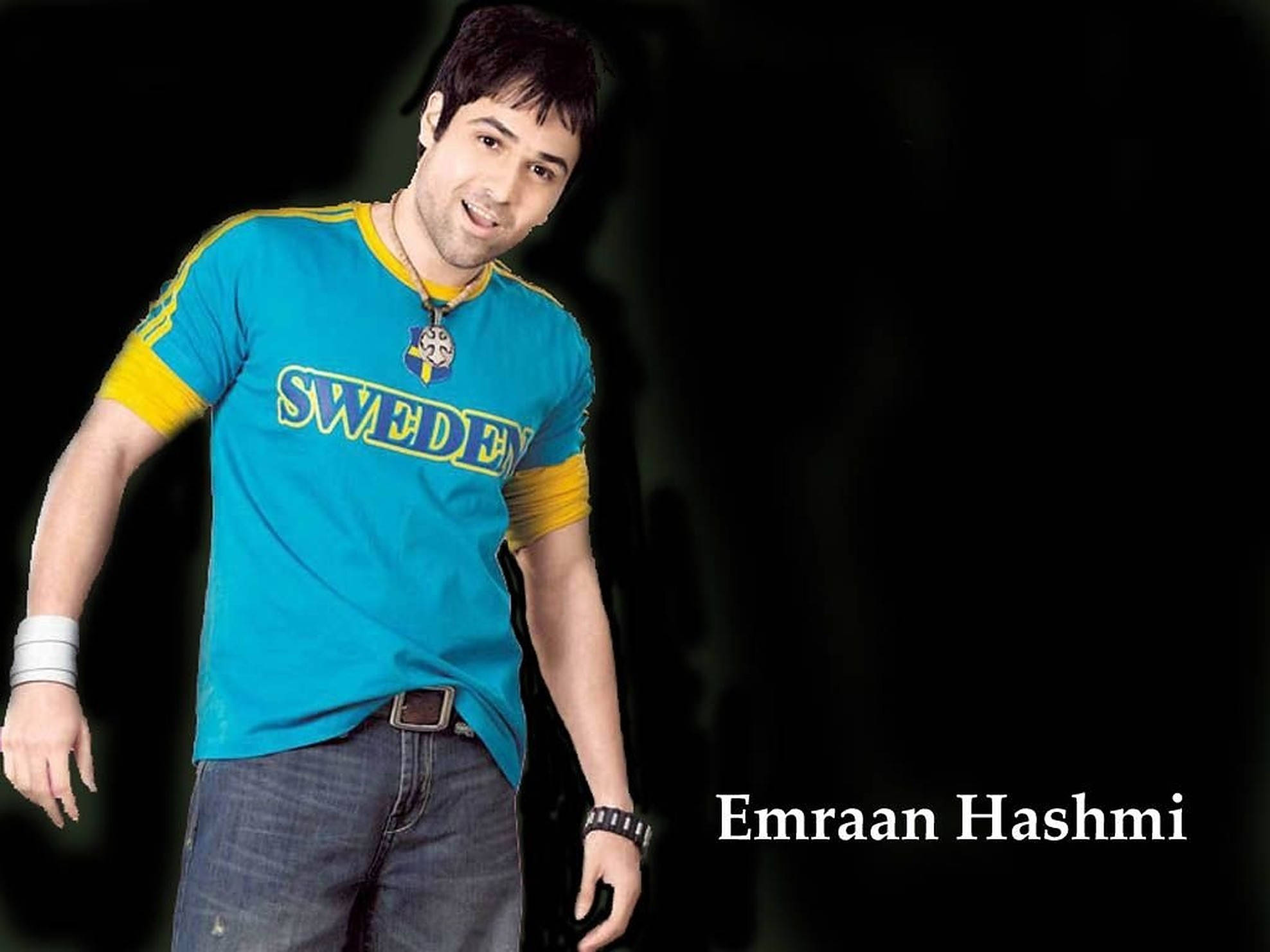 Emraan Hashmi 桌布 壁紙 — 一名身穿藍黃色瑞典T恤的男子在深色背景下自信地微笑。