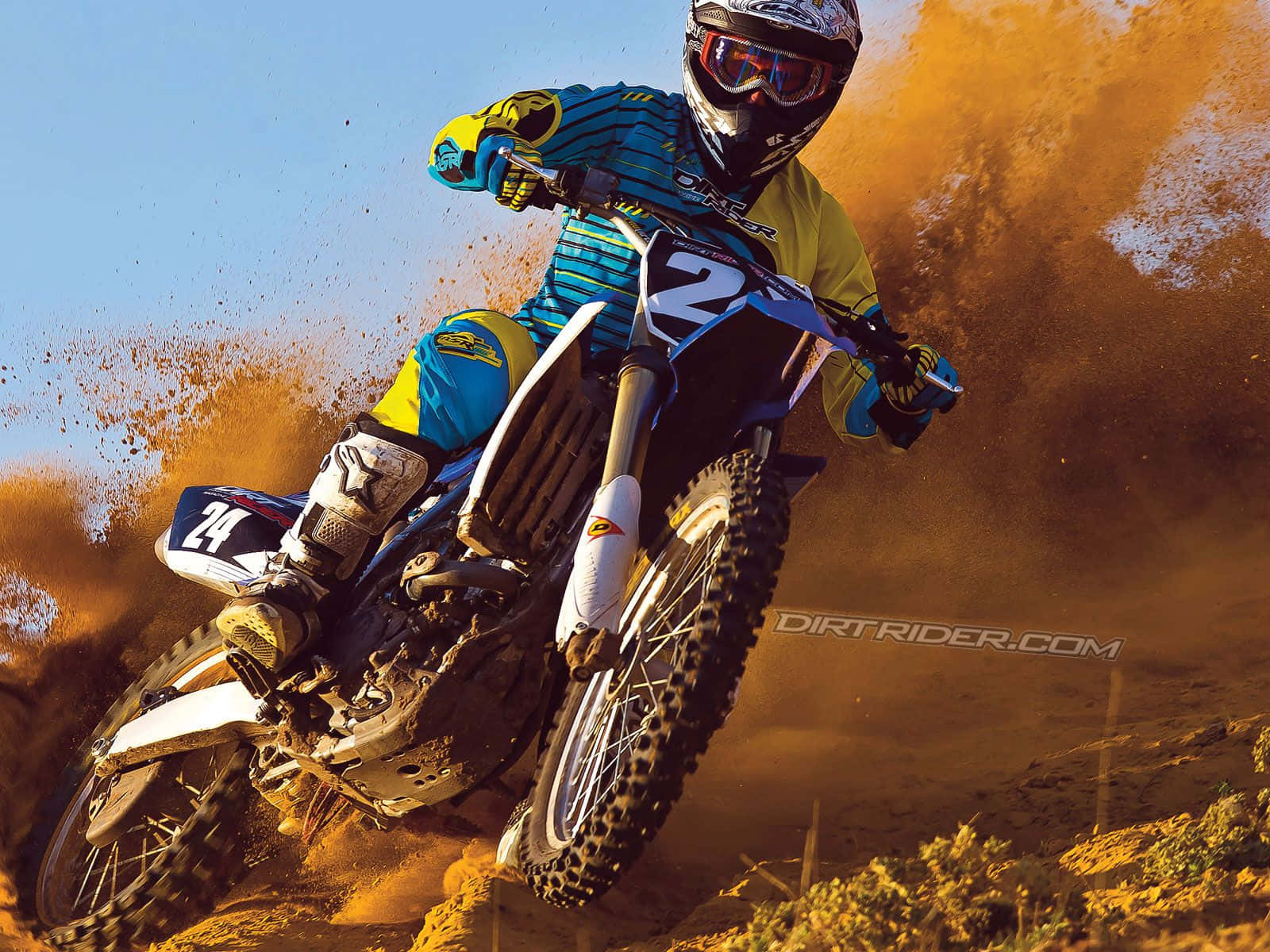 Honda Dirt Bike 桌布 壁紙 — 一名身穿藍黃色裝備的越野摩托車手在晴朗藍天下傾斜入彎，揚起橙色塵土。
