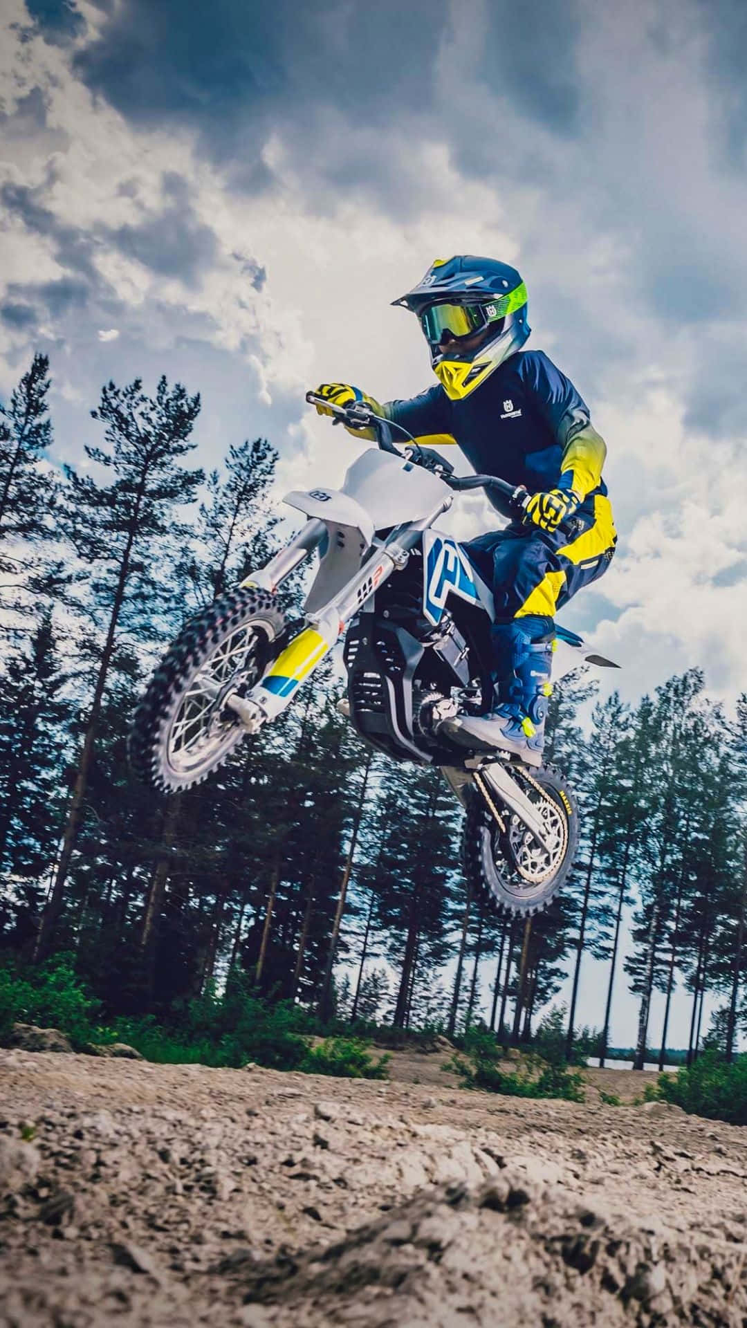 Dirtbike 桌布 壁紙 — 一名身穿藍黃色裝備的越野機車手，在陰天之下，騎著白色越野機車飛越森林小徑上空。