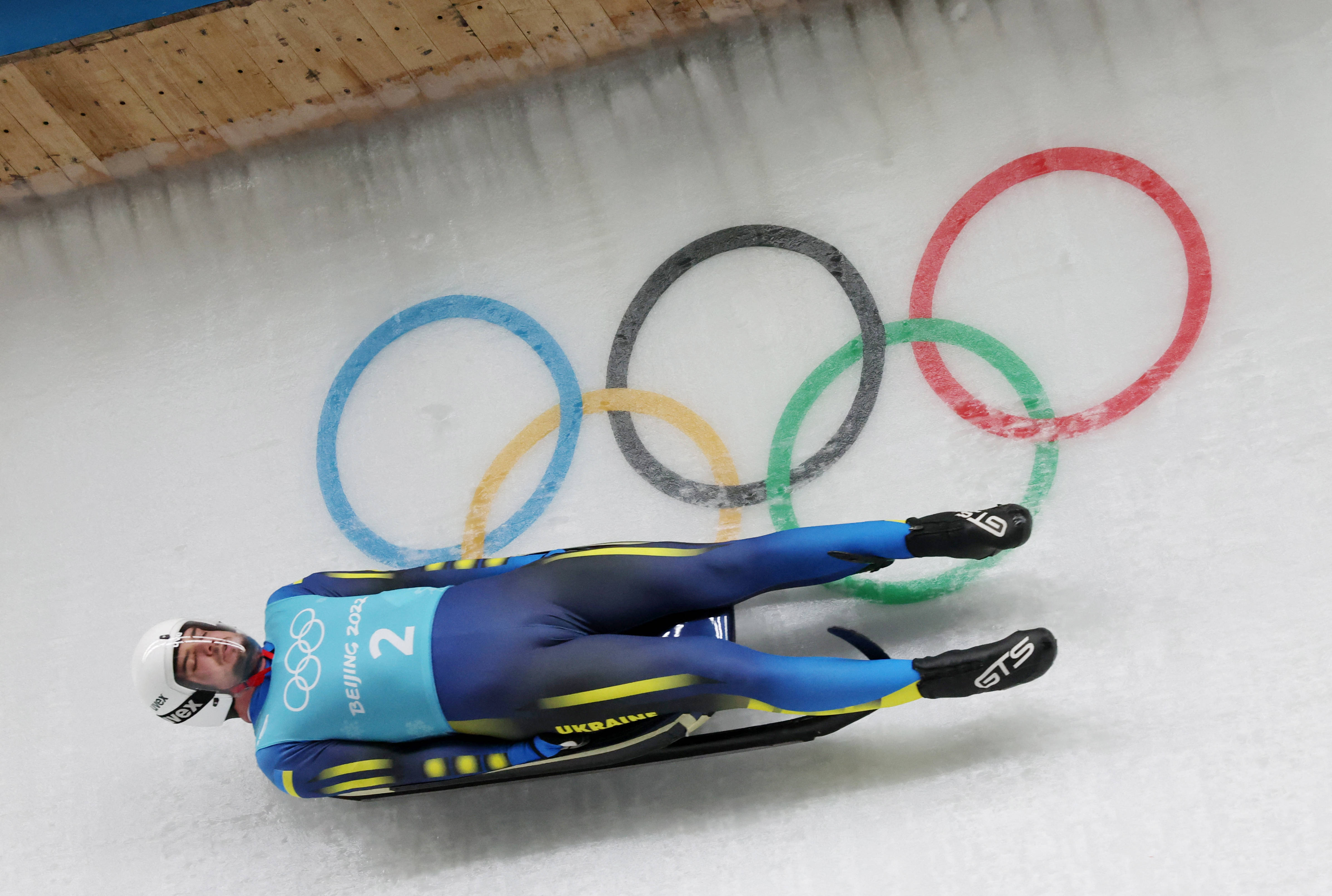 luge 桌布 壁紙 — 一名身穿藍黃色裝備的雪橇運動員在冰道上飛馳，身後冰面上可見奧運五環。