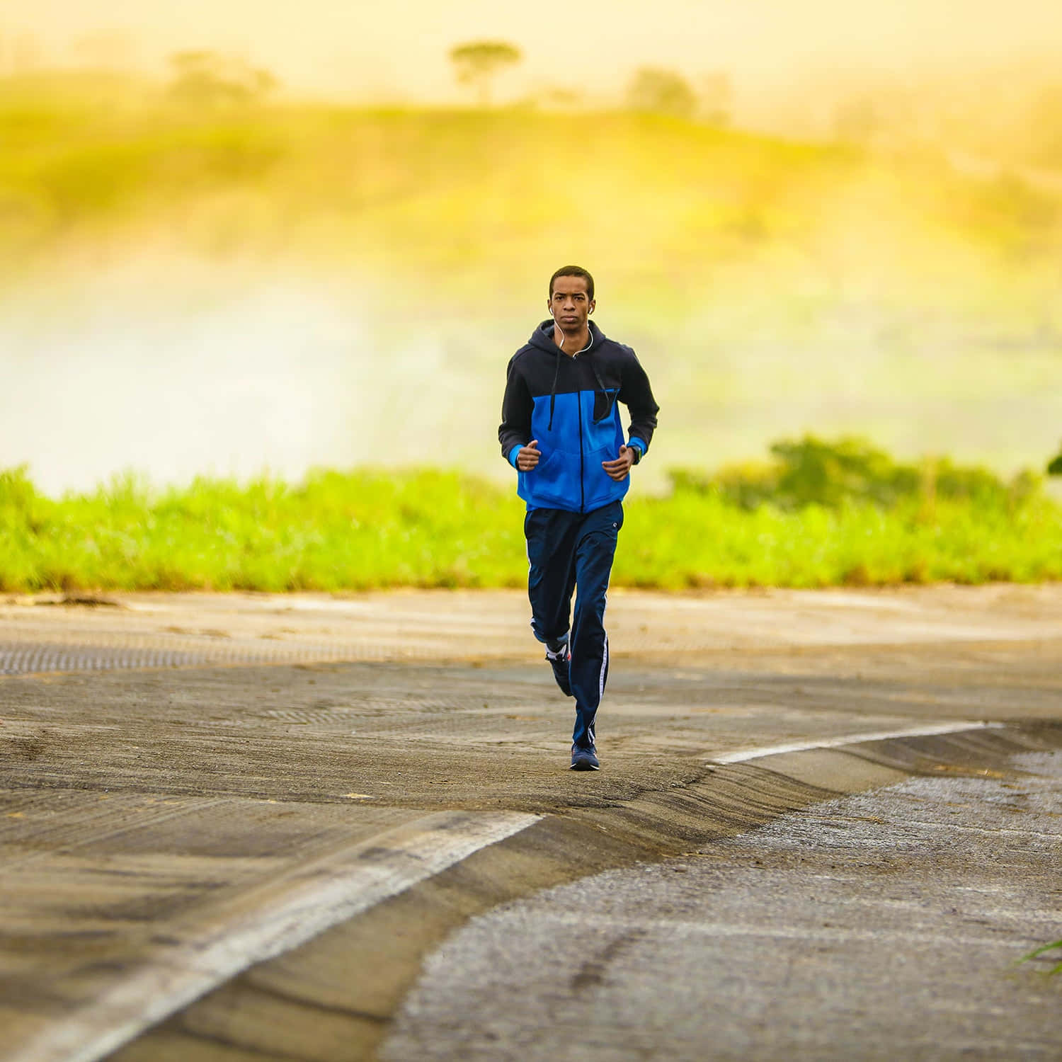 Running 桌布 壁紙 — 一名身穿藍黑夾克的男子在鋪砌的道路上奔跑，路旁有綠草和朦朧的山丘，天空溫暖金黃。
