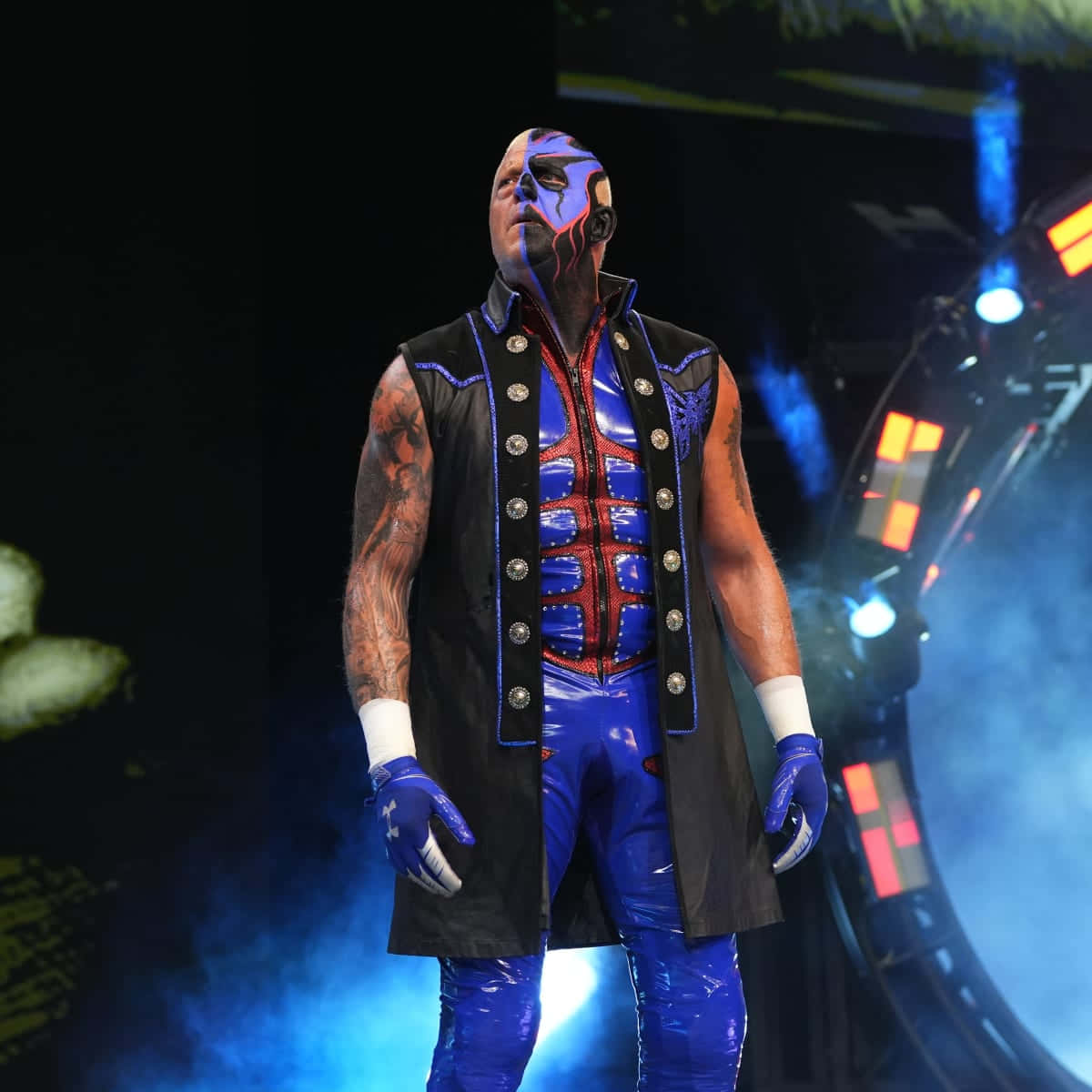WWE 桌布 壁紙 — 一名身穿藍黑服裝、臉上塗有顏料的摔角手，在煙霧繚繞的舞台燈光下戲劇性地站立。
