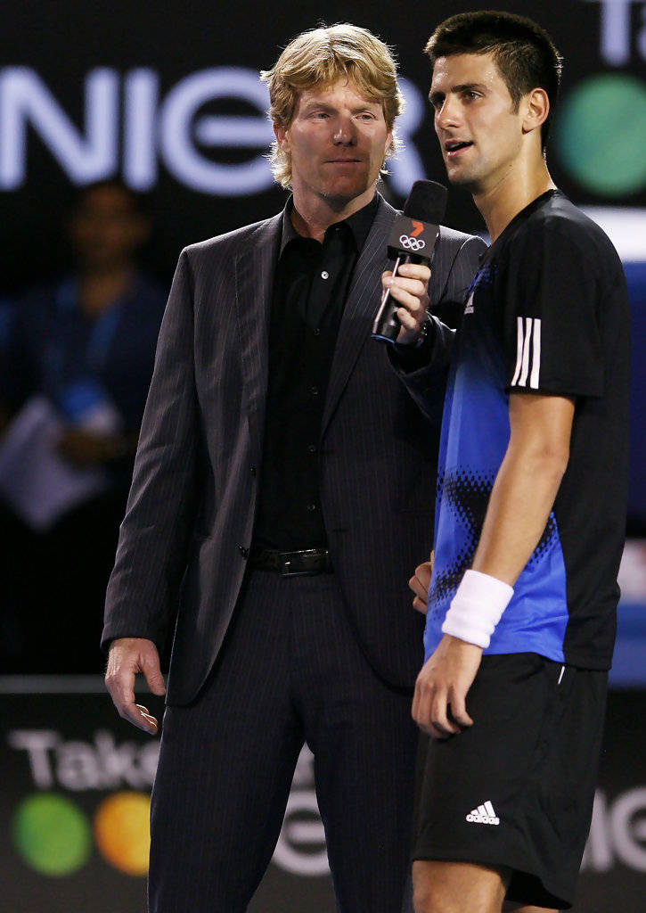 Tennis 桌布 壁紙 — 一名身穿藍黑色運動服的男網球選手，站在一位身穿西裝、手持麥克風的採訪者旁邊，背景是燈光明亮的體育舞台。