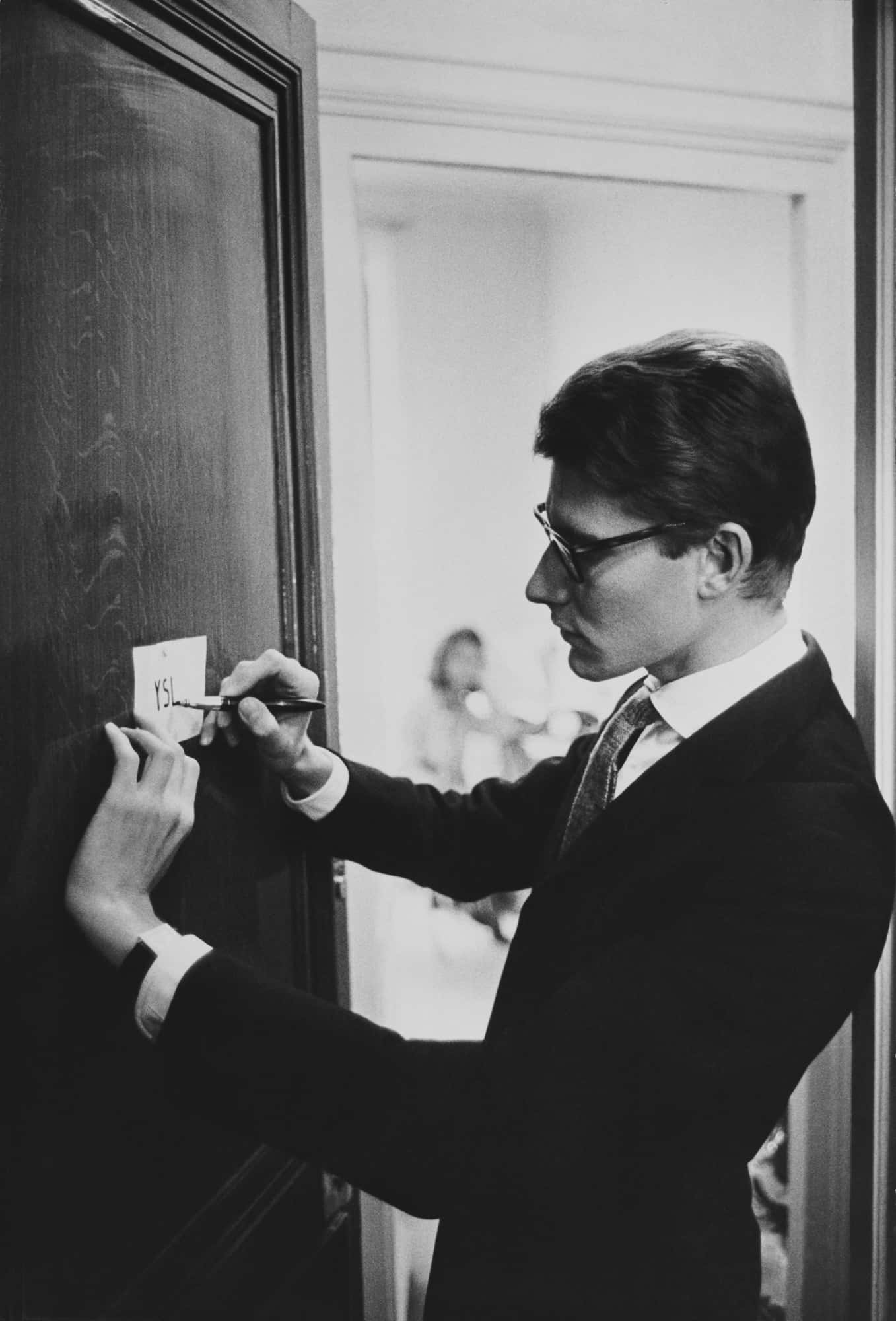 YSL 桌布 壁紙 — 一名身穿西裝、戴眼鏡的男子，在一張黑白照片中，於深色木門上的便利貼寫上「YES」。
