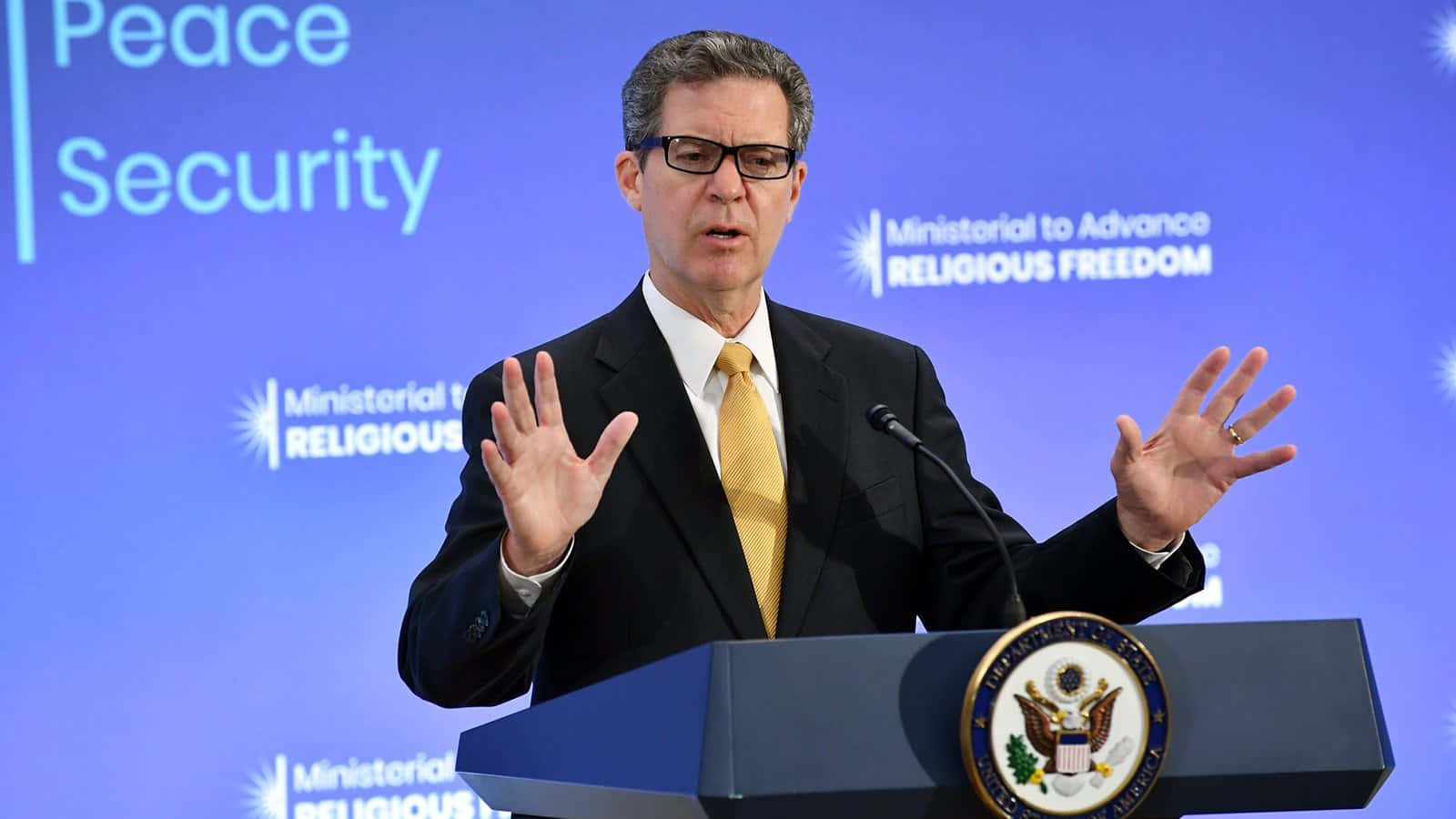 政治 桌布 壁紙 — 一名身穿西裝、戴眼鏡的男子，在帶有美國國徽和「Ministerial to Advance Religious Freedom」背景的講台上發言並做手勢。