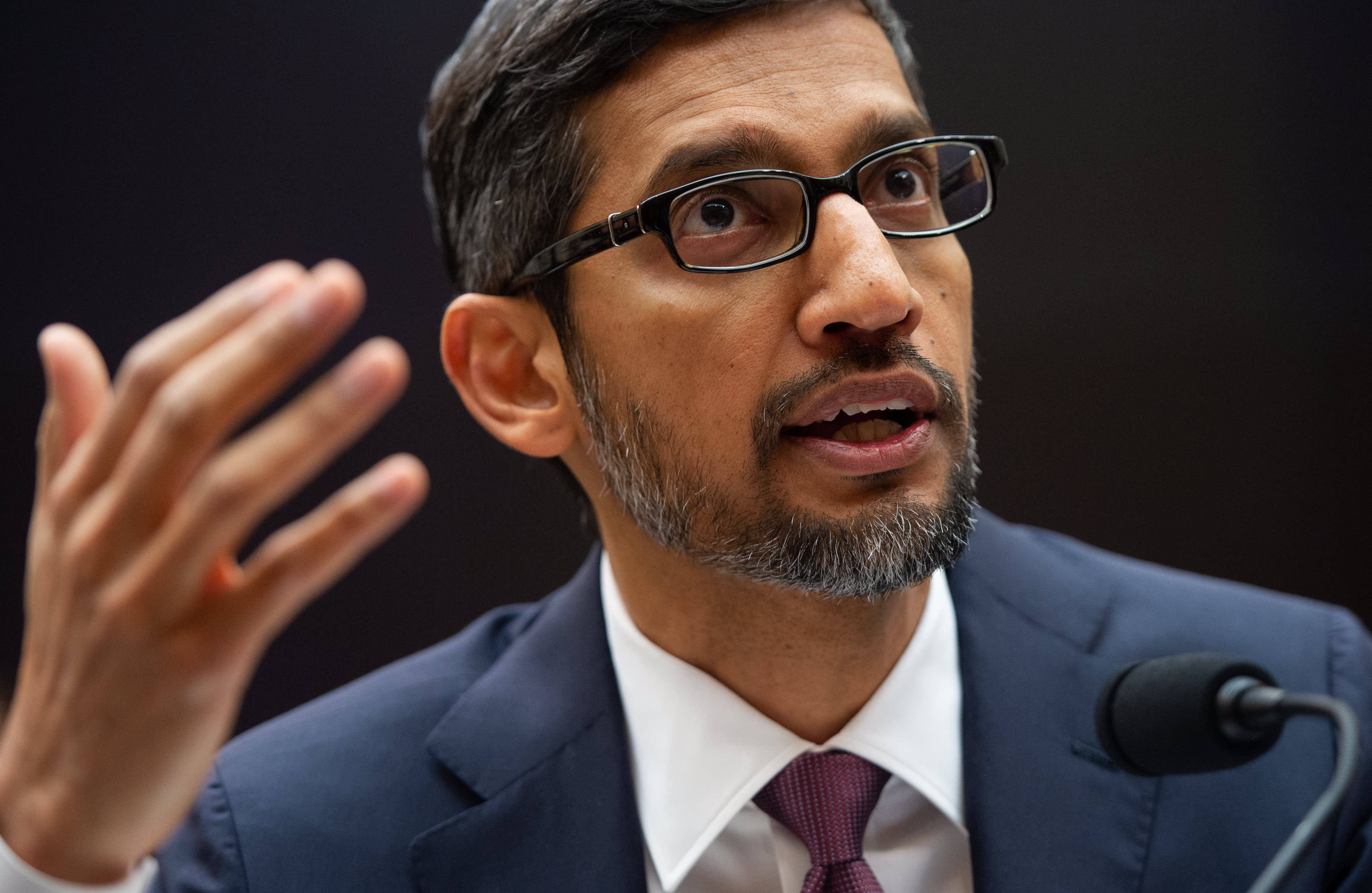 google 桌布 壁紙 — 一名身穿西裝、戴眼鏡的男子在深色背景下對著麥克風說話時做手勢的特寫。