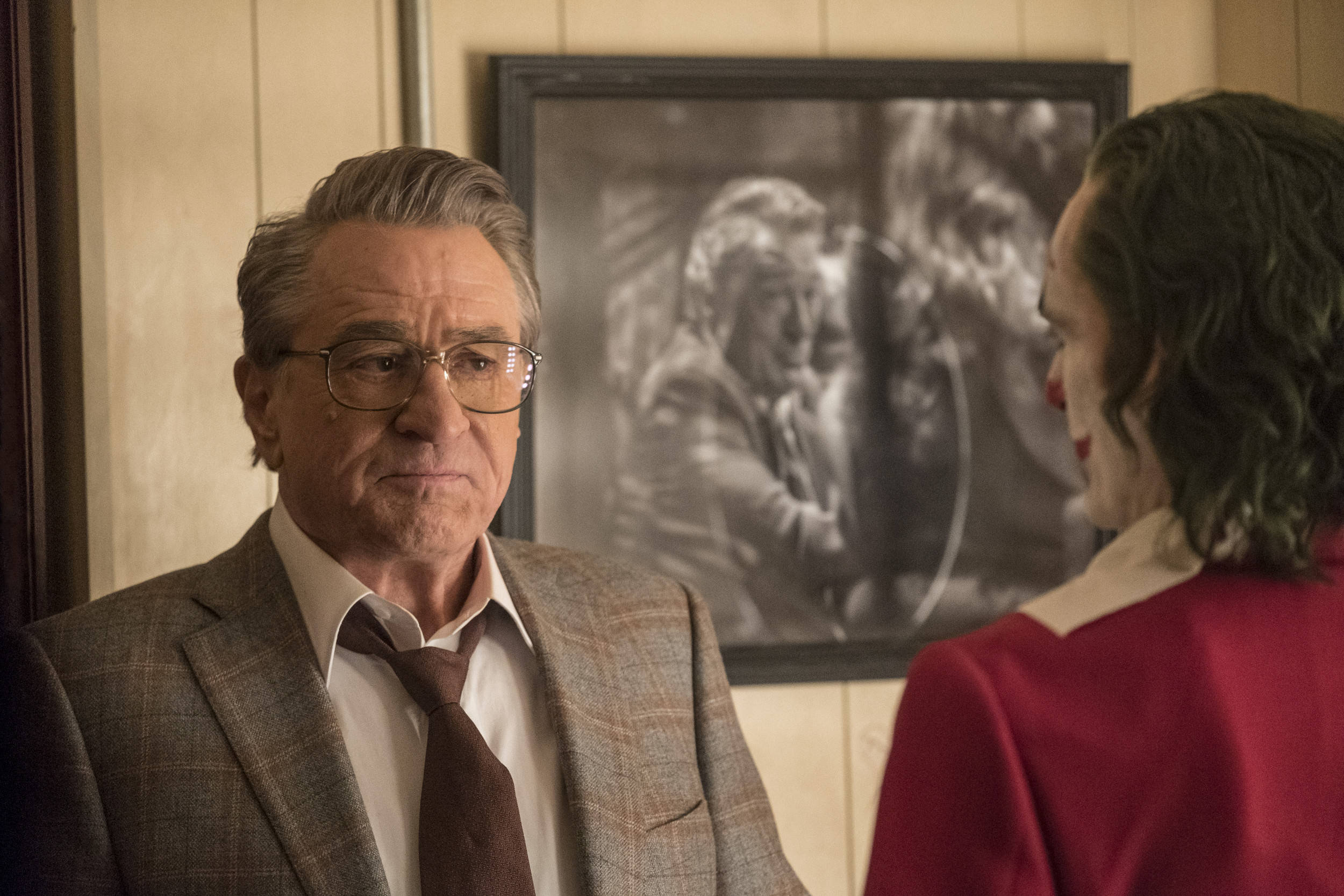 Robert De Niro 桌布 壁紙 — 一名身穿西裝、戴眼鏡的男子在燈光溫暖的房間裡，專注地凝視著一位身穿紅色外套、白色領子的人。