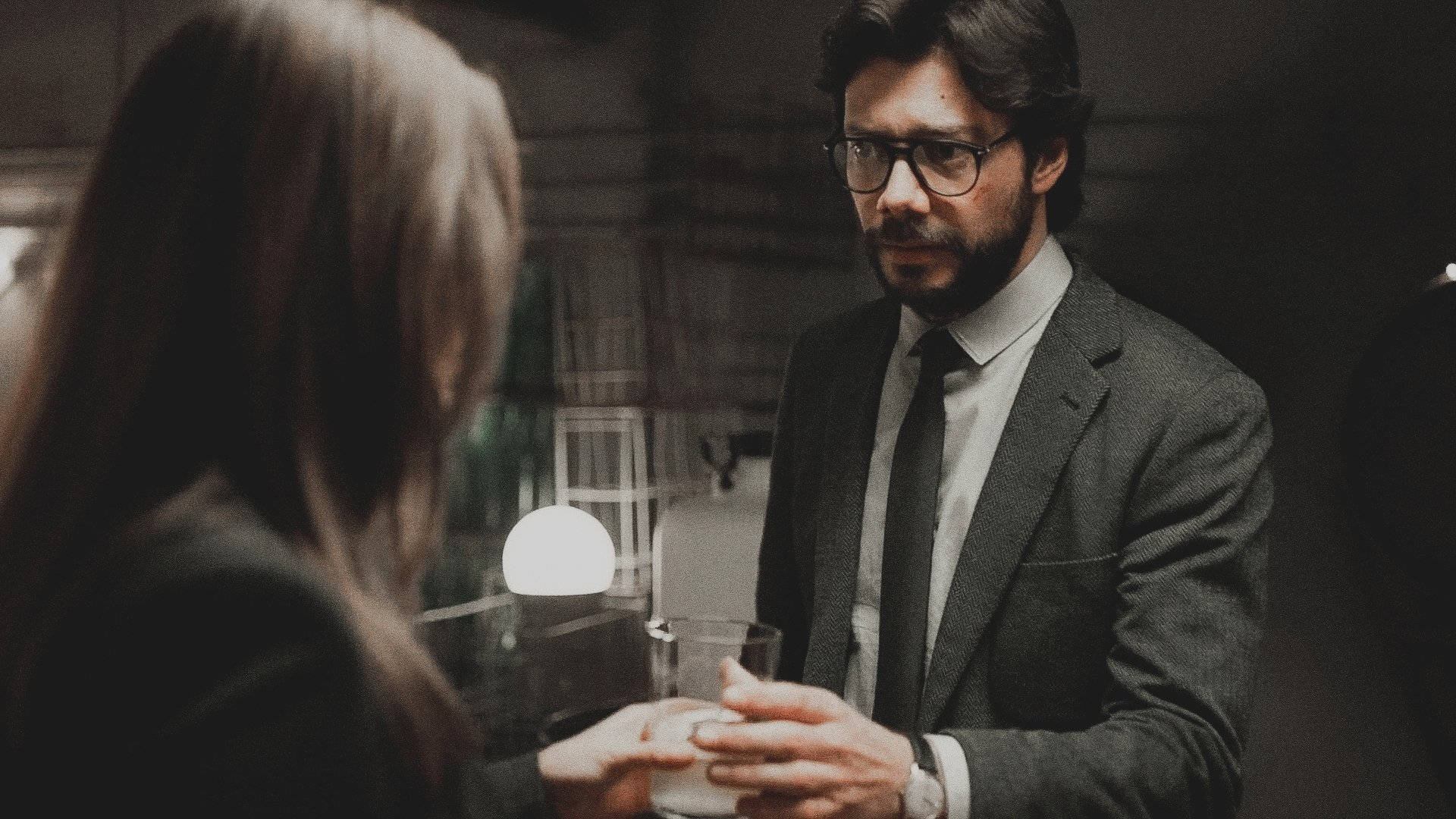Professor 桌布 壁紙 — 一名身穿西裝、戴眼鏡的男子手持酒杯，在燈光昏暗的現代室內與一名女子進行嚴肅對話。