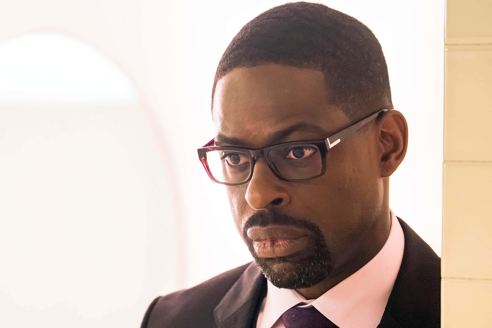 Sterling K Brown 桌布 壁紙 — 一名身穿西裝、戴著眼鏡的男子，在明亮的背景下以嚴肅的表情專注地凝視。
