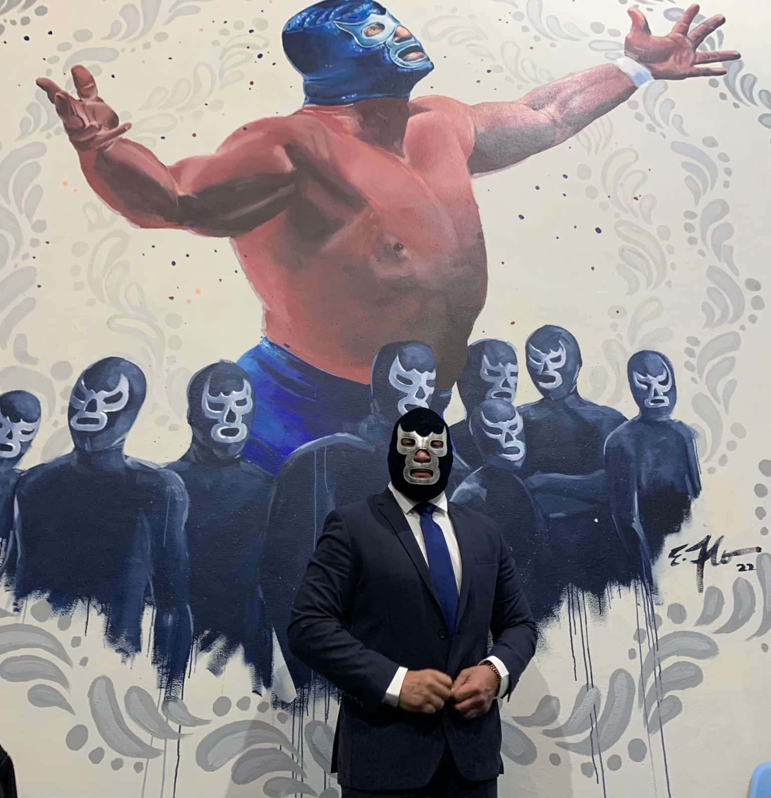 graphic design 桌布 壁紙 — 一名身穿西裝、戴著自由摔角面具的男子站在一幅肌肉發達的蒙面摔角手壁畫前，該摔角手雙臂張開。