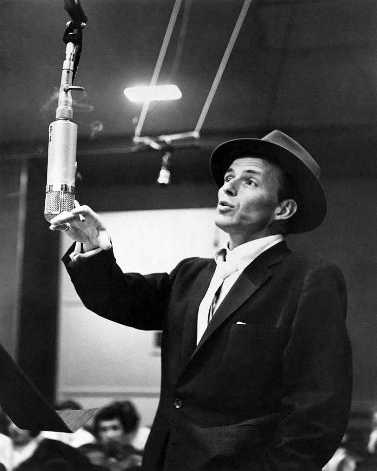 Sinatra 桌布 壁紙 — 一名身穿西裝、戴著費多拉帽的男子在錄音室中手持復古麥克風，頭頂有明亮的燈光。
