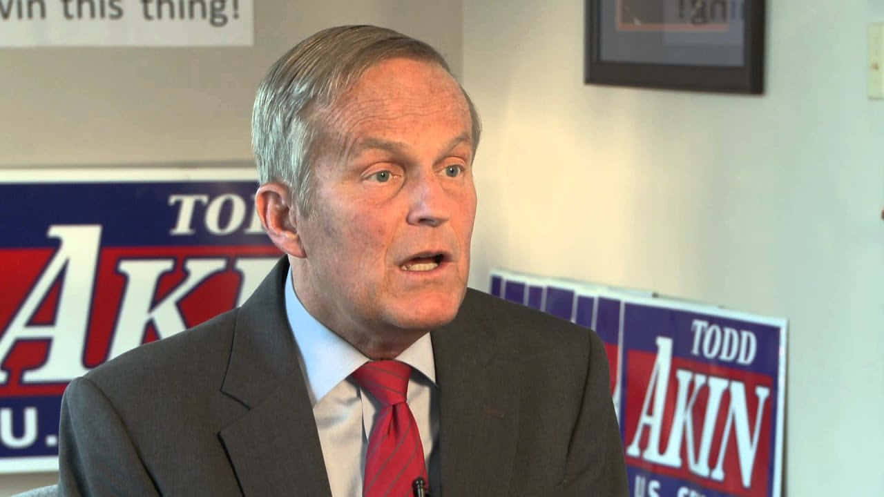 Election 桌布 壁紙 — 一名身穿西裝、打紅色領帶的男子在 Todd Akin 的競選標誌前認真發言。