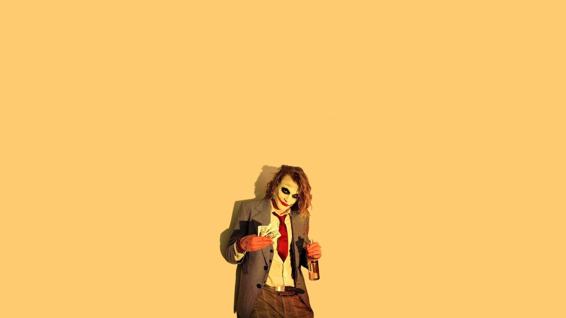 Heath Ledger Joker 桌布 壁紙 — 一名身穿西裝、紅色領帶、白色臉部彩繪和紅色嘴唇的男子，在純黃色背景前拿著金錢和一個瓶子。