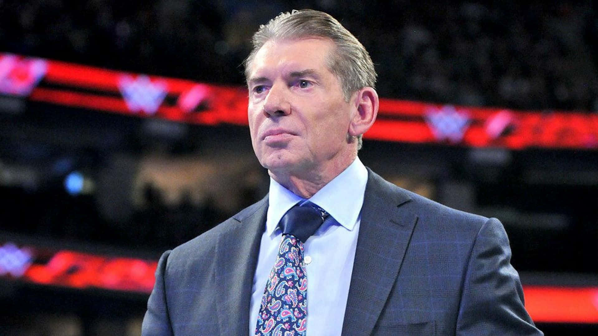 WWE 桌布 壁紙 — 一名身穿西裝和佩斯利領帶的男子，在身後有紅色發光燈光的昏暗場館中莊嚴地站立。