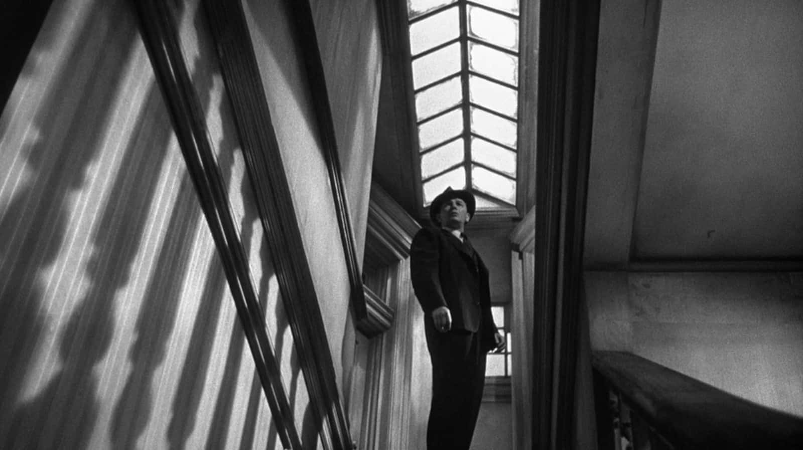 Film Noir 桌布 壁紙 — 一名身穿西裝和帽子的男子站在樓梯上，被高窗投射的戲劇性條紋光線所籠罩。