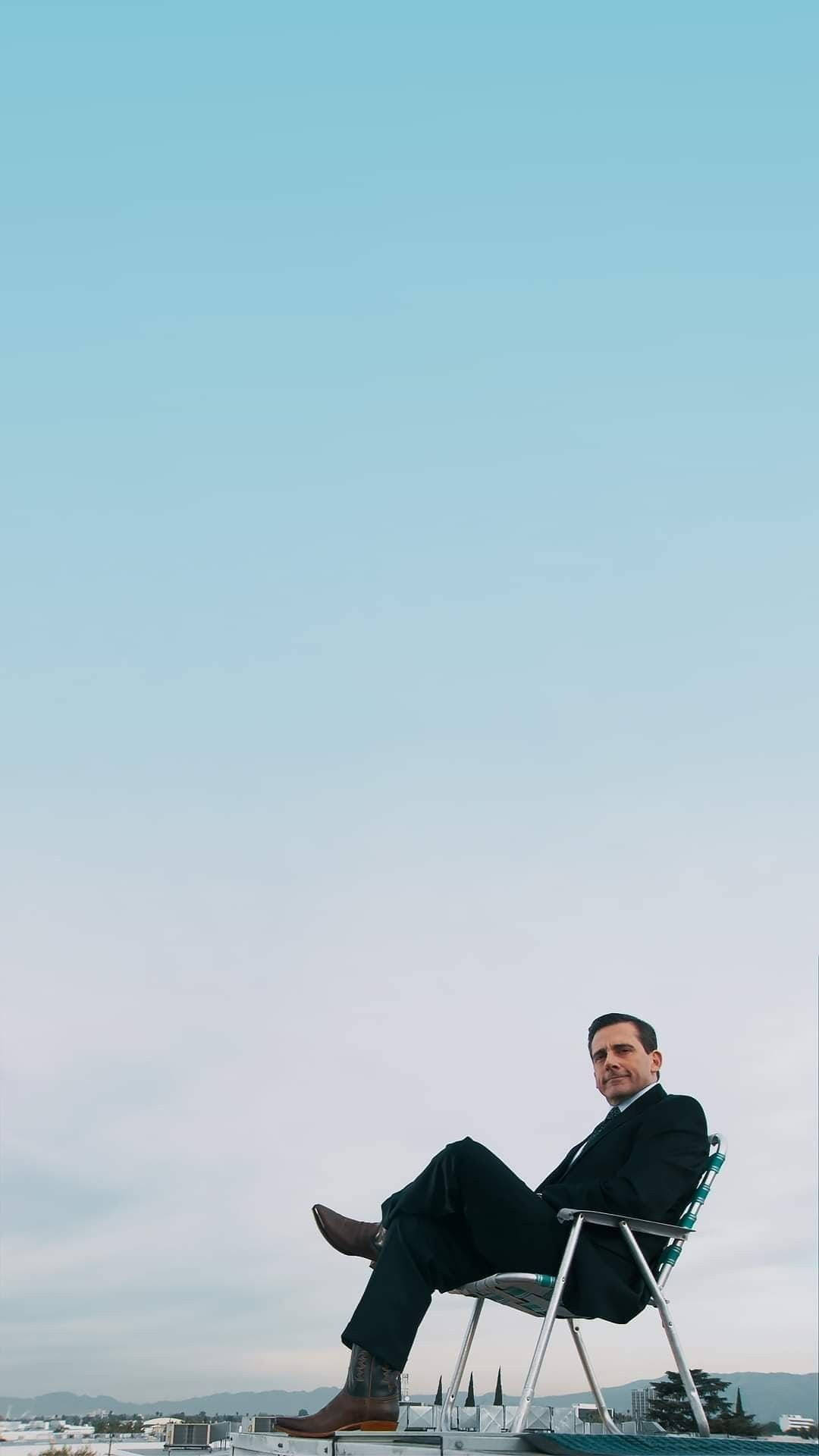 Michael Scott 桌布 壁紙 — 一名身穿西裝和牛仔靴的男子，在廣闊的淡藍色天空下，放鬆地坐在屋頂的椅子上。
