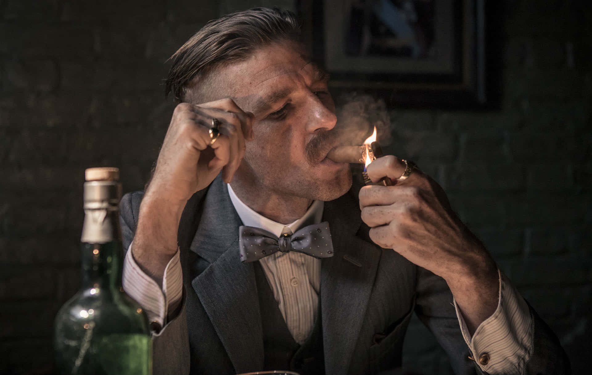 西裝男子在昏暗磚房點雪茄，旁有瓶子 — Peaky Blinders 壁紙