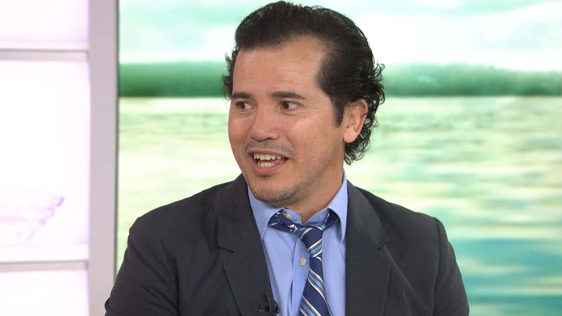 John Leguizamo 桌布 壁紙 — 一名身穿西裝打領帶的男子在模糊水背景的電視佈景中微笑說話。