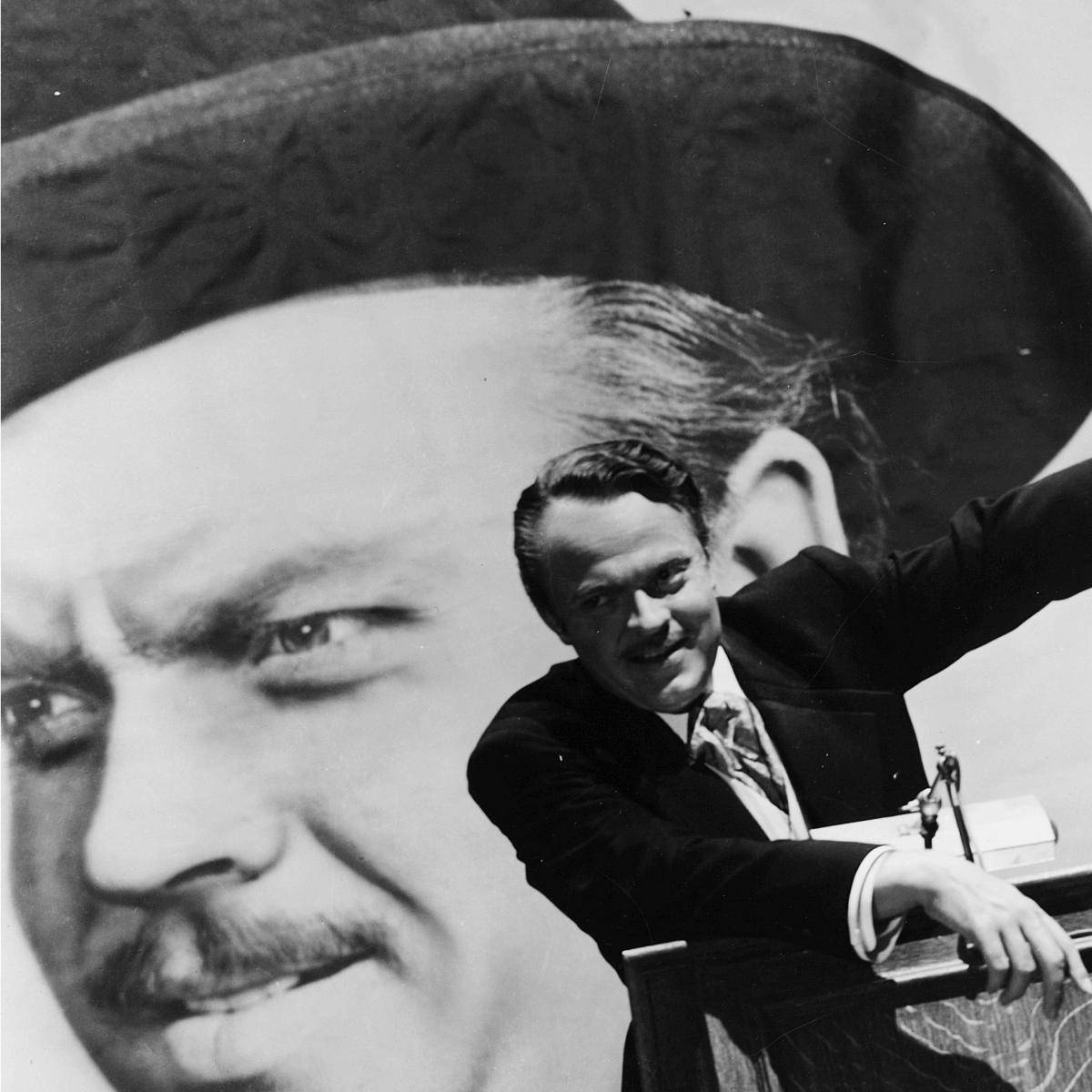 Citizen Kane 桌布 壁紙 — 一名身穿西裝打領帶的男子站在講台上，面對一幅巨大的黑白肖像，肖像中是一名留鬍子戴帽子的男子，他面帶微笑。