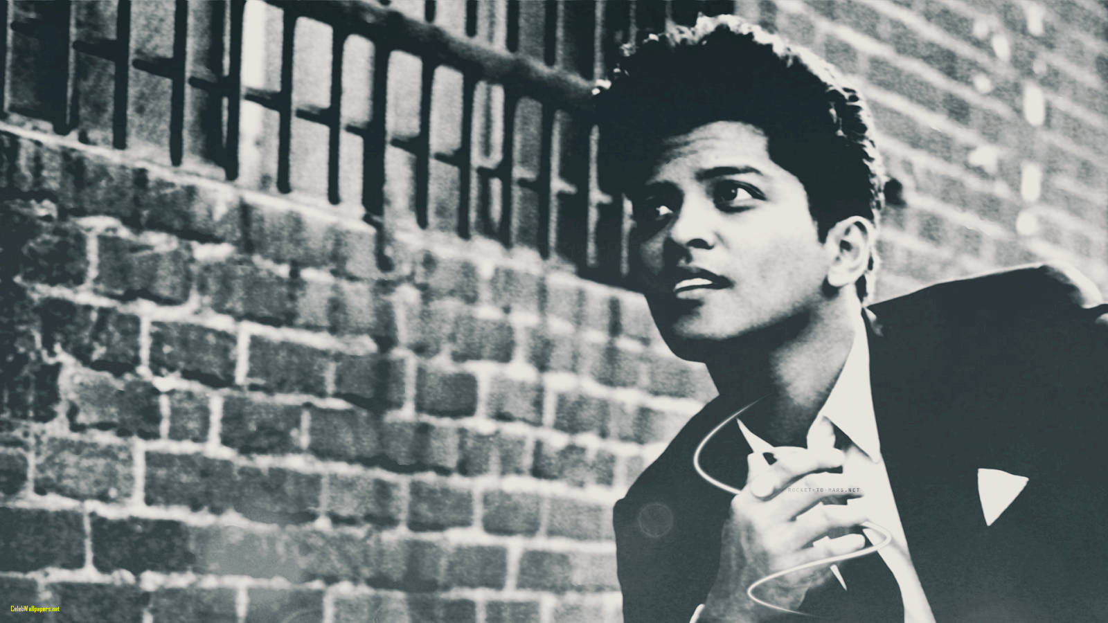 Bruno Mars 桌布 壁紙 — 一名身穿西裝的年輕男子靠著有金屬欄杆的磚牆，抬頭仰望，營造出黑白肖像的憂鬱氛圍。
