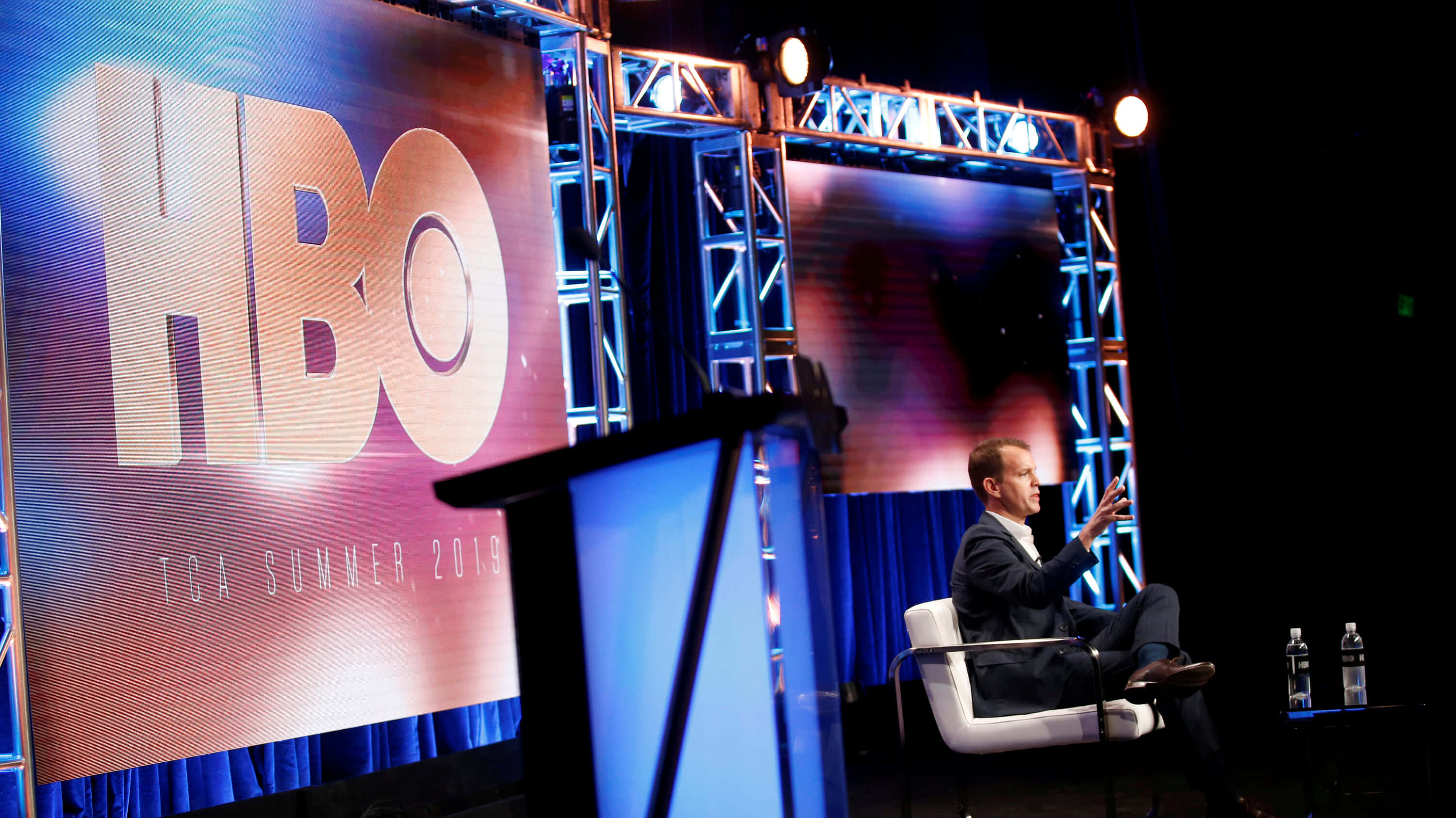 HBO 桌布 壁紙 — 一名身穿西裝的男子在HBO TCA Summer 2019活動的舞台上，在明亮的舞台燈光下坐著做手勢。