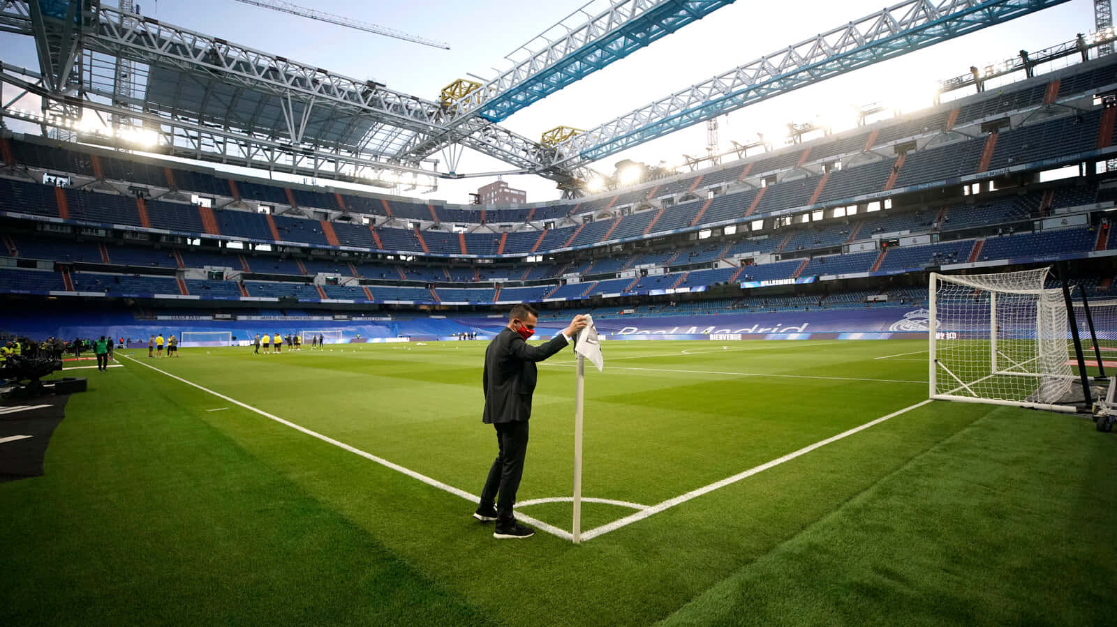 Soccer Stadium 桌布 壁紙 — 一名身穿西裝的男子在一個空無一人的足球場上擦拭角球旗，球場有藍色座位和明亮的綠色草坪，在日光下。