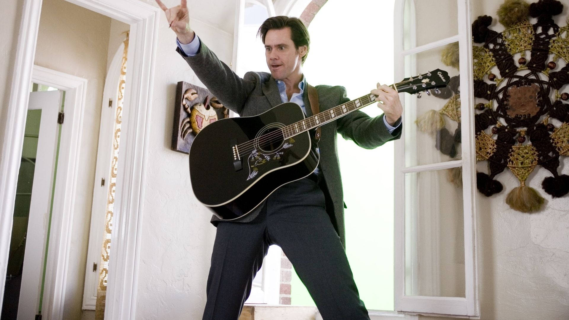 Jim Carrey 桌布 壁紙 — 一名身穿西裝的男子在光線明亮的走廊中充滿活力地彈奏黑色木吉他，同時向上指。
